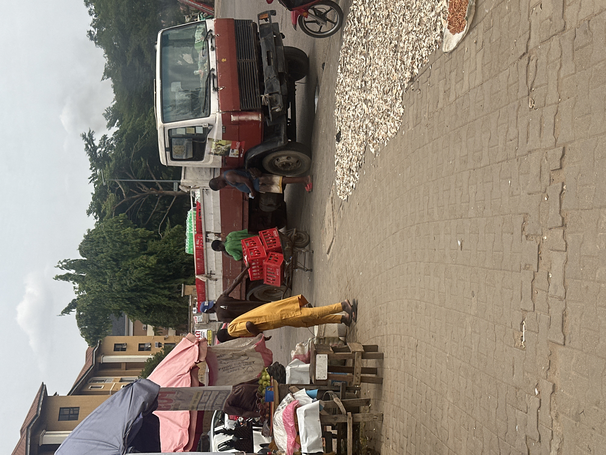 The image depicts a street scene in Abuja, Nigeria. A red and white truck is parked on the side of the road. A man is standing near the truck, and two other men are carrying crates, possibly of beverages, from the truck. A pile of small, white objects, likely dried goods, is laid out on a sheet on the pavement next to the truck. In the foreground, a man in a yellow outfit appears to be examining goods displayed on a wooden structure near a market stall covered with umbrellas and various items for sale. Buildings and trees are visible in the background, suggesting an urban or semi-urban environment.