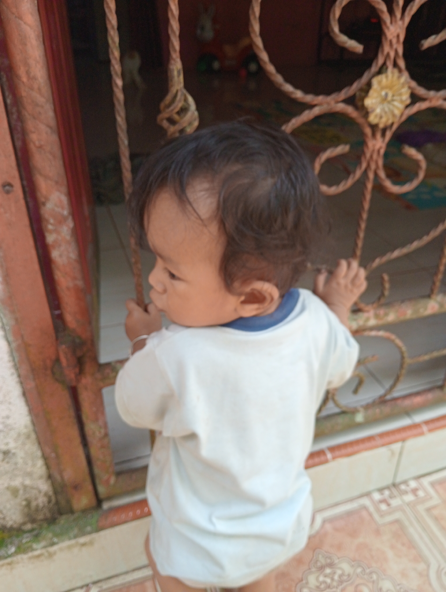 A young child, likely a toddler, stands facing away from the camera, peering through a decorative metal gate. The child is wearing a white t-shirt with a blue collar and a thin silver bracelet on their left wrist. Their dark hair is slightly messy. The child's right hand is gripping a horizontal bar of the gate, and their left hand is also near the gate. The gate appears to be made of twisted metal with a decorative floral element. Behind the gate, the background is blurred but suggests an indoor or covered space with colorful floor tiles and what appears to be toys or objects on the floor. The foreground shows a textured tiled floor and the base of a reddish-brown structure, possibly a wall or pillar. The overall scene suggests a residential setting, potentially in Campago Barat, Indonesia, where the child is exploring or observing their surroundings.