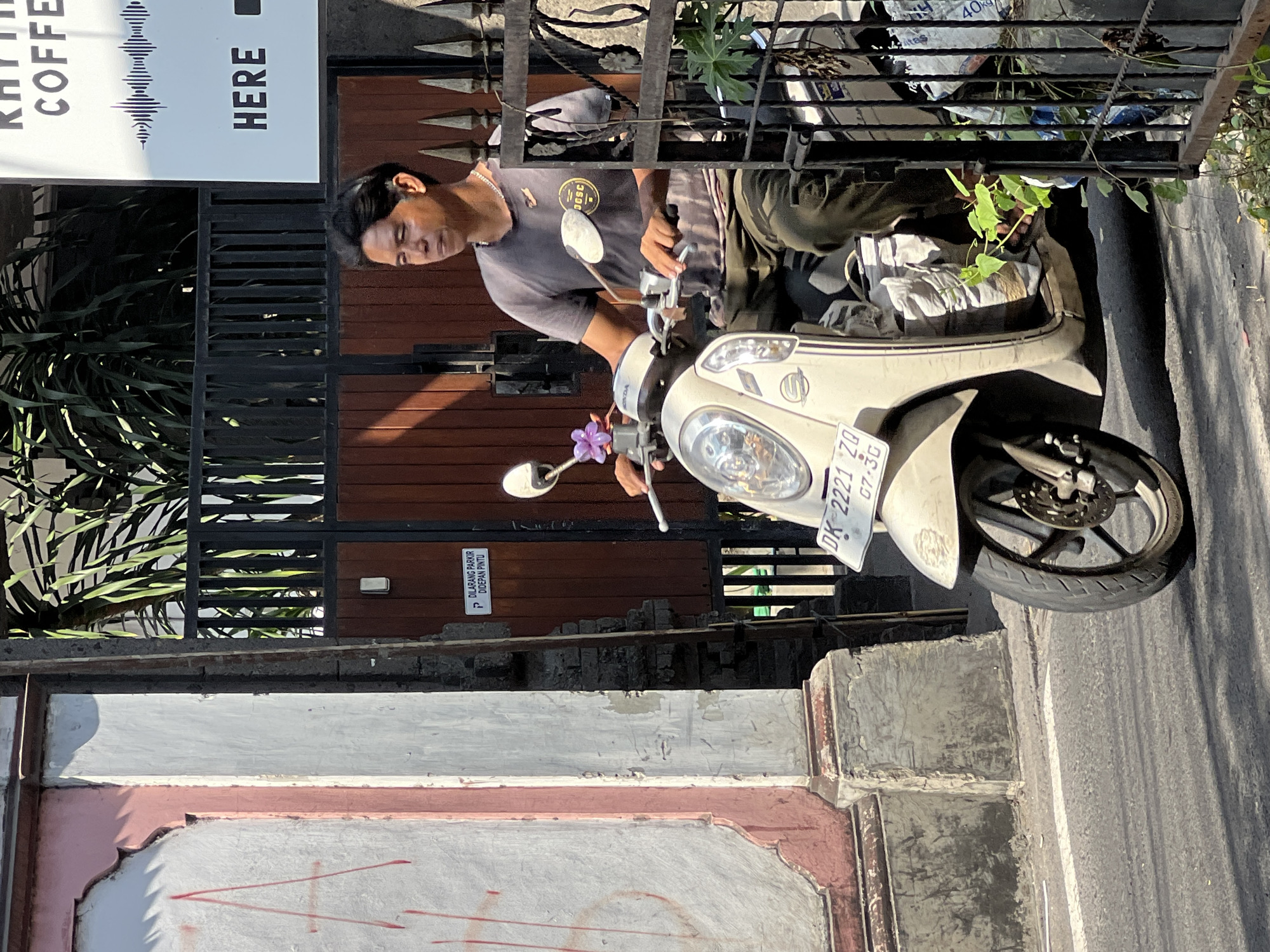 A man rides a light-colored scooter down a street in Kuta, Indonesia. He wears a dark t-shirt with a circular logo and dark pants. The scooter has a purple flower attached to the handlebars. Behind the scooter, a wooden gate and fence are visible, along with a sign that reads "RHYTHM COFFEE" and "HERE." To the left of the scooter, a textured pink and white wall is present. The street is paved and appears to be a public road. Sunlight casts shadows on the ground.