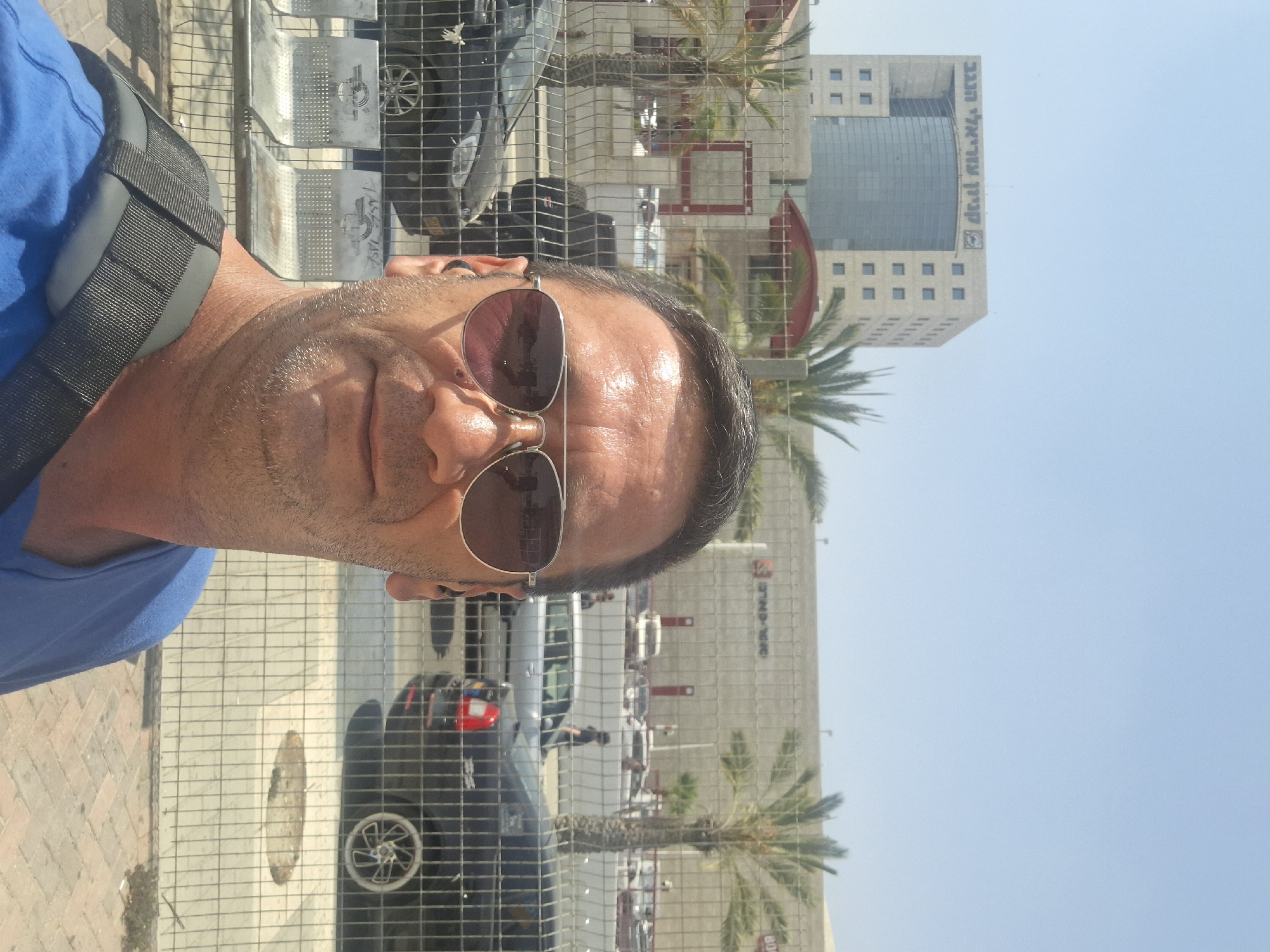 A man wearing sunglasses and a blue shirt takes a selfie outdoors. Behind him, a building with "קניון עזריאלי הנגב" (Azrieli Negev Mall) written on it stands tall. Palm trees are visible in the background, and cars are parked in a lot. A fence separates the man from the background scene. The man appears to be near the Azrieli Negev Mall in Be'er Sheva, Israel.