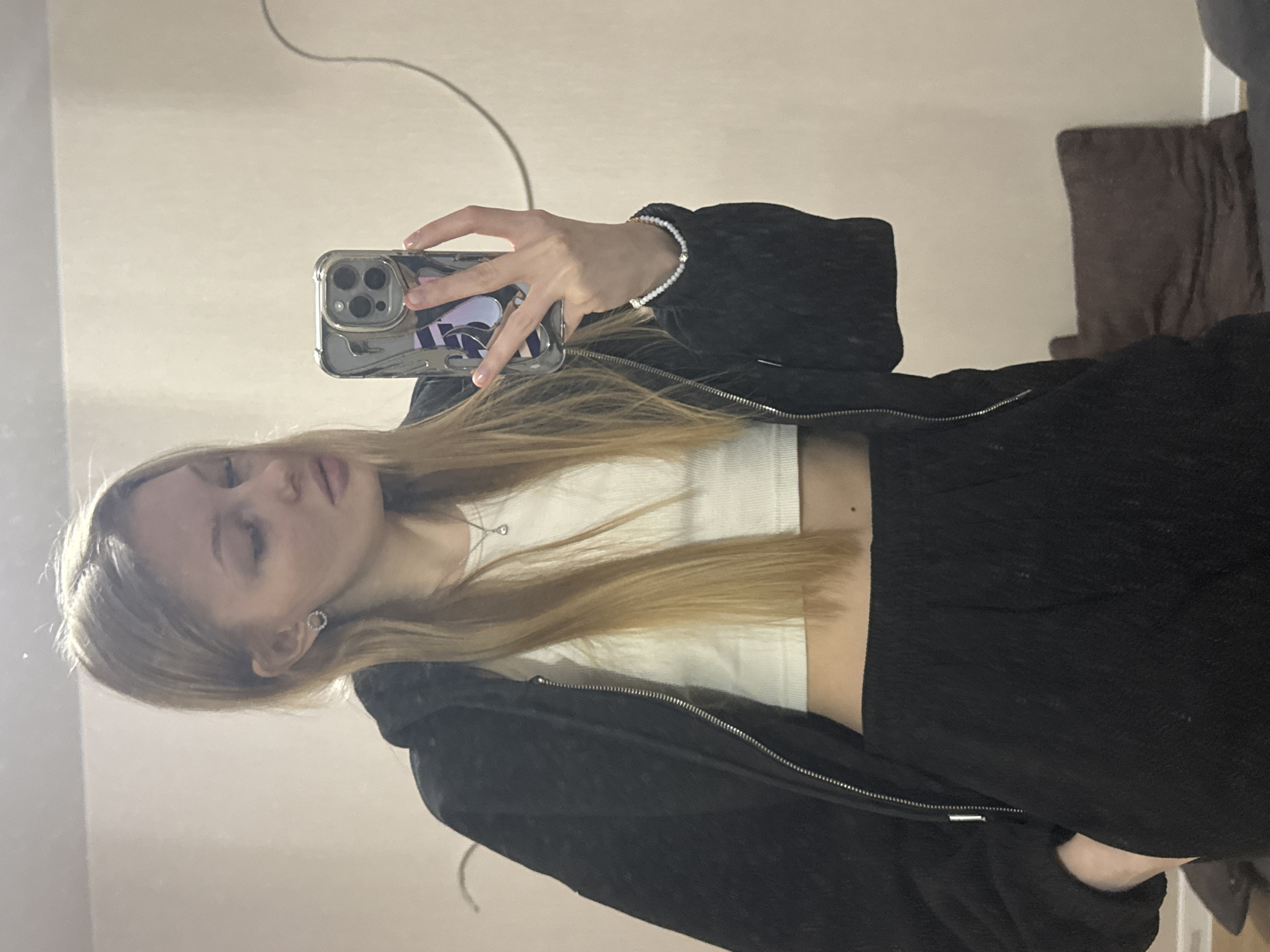 The image, taken in Perm, Russia, depicts a young woman capturing a mirror selfie indoors. She appears to be in her late teens or early twenties, characterized by long, straight, blonde or light brown hair that cascades over her shoulders and chest. Her attire includes a white ribbed crop top, a black textured zip-up hoodie or jacket, and dark-colored bottoms, likely pants or shorts.
The woman holds a smartphone, identifiable as an iPhone (possibly a Pro Max model due to its triple camera array), in a clear protective case with her right hand. On her right wrist, she wears a white beaded bracelet adorned with small golden charms. A delicate silver necklace with a tiny, clear pendant is visible around her neck. Her left ear features a small, circular silver earring, and a mole is visible on the left side of her neck.
Her face is captured in profile, with her eyes cast downwards or closed, conveying a serene or pensive expression. The background, which is the mirror's reflection of the room, consists mainly of a plain, light-colored wall. Subtle details in the reflection include what appears to be a dark brown curtain or fabric on the right and possibly a dark piece of furniture or object on the left. A faint cable or wire is visible across the upper portion of the background. The lighting is soft and even, consistent with an indoor setting. There is no visible text within the image.