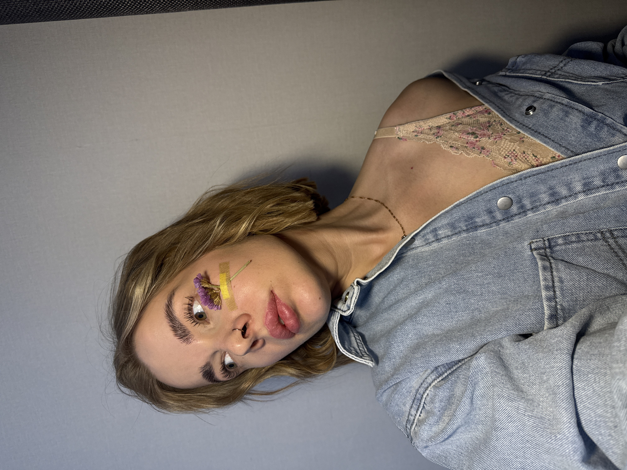 A woman is seen in a close-up shot against a plain gray background. She has light brown hair and a flower taped to her face above her right eye. She is wearing a denim jacket, which is partially unbuttoned, revealing a lace bralette. A gold necklace is visible around her neck. The setting and location, Chaikovsky, Russia, are not identifiable from the image.