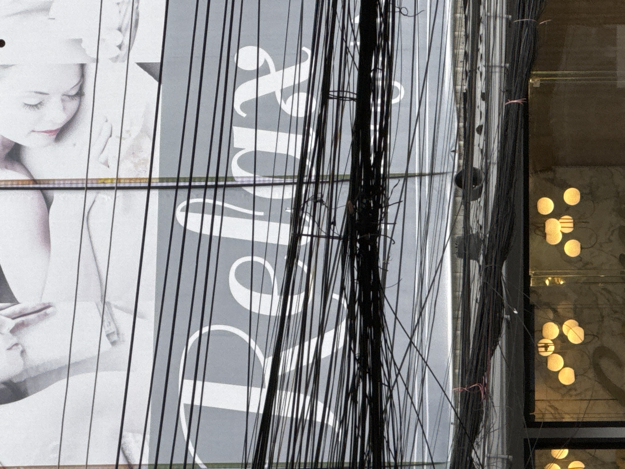 The image shows a close-up view of a grey sign with white lettering that reads "Relax". The sign is partially obscured by a dense tangle of black electrical wires, which are strung across the foreground. Above the sign, a portion of a larger billboard is visible, featuring black and white images of a person's head and torso, with a hand resting on their chest. The billboard has a light grey background. Below the sign and wires, a section of a building facade is visible, with large glass windows reflecting warm, yellow circular lights that appear to be hanging fixtures. The scene appears to be outdoors, likely in an urban or commercial area.