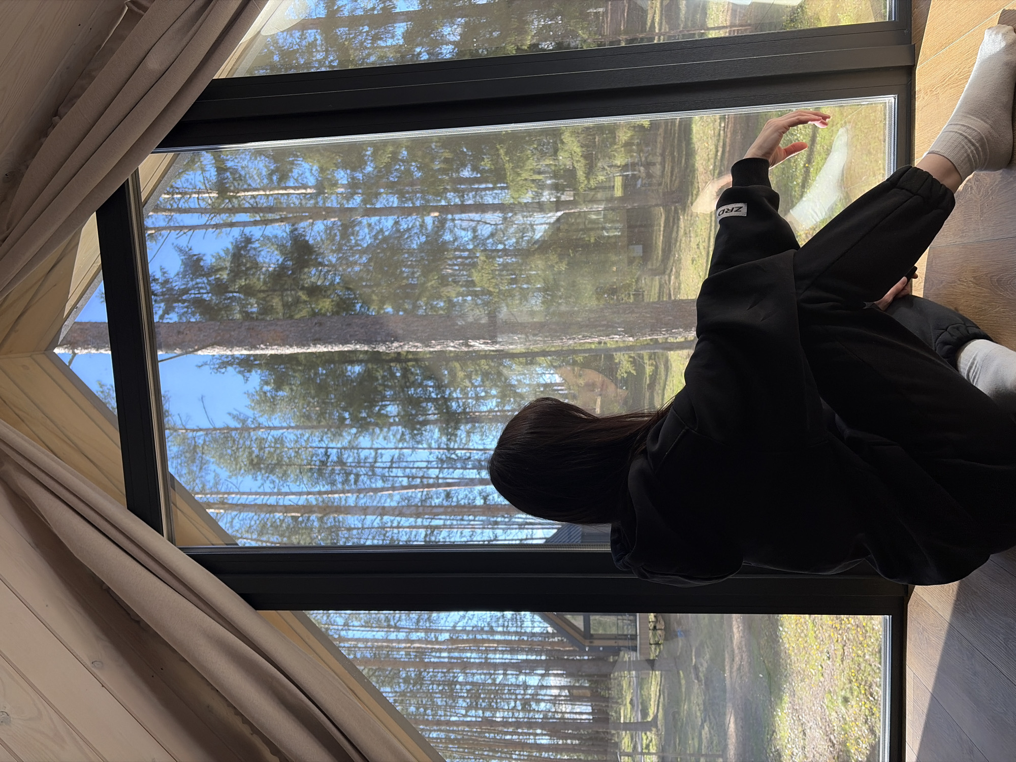 An image captures a person viewed from the back and side, sitting on a dark wooden floor beside a large, dark-framed window. The person is wearing a black hoodie with "ZRD" in white text on the left sleeve, black sweatpants, and light-colored socks. Their right hand is slightly raised, fingers splayed, near the windowpane, while their left leg is bent and the right leg is extended along the floor.
Through the window, a bright, clear blue sky and a dense pine forest are visible. Tall, slender pine tree trunks dominate the outdoor scene, with green foliage at their tops. The ground outside appears grassy with some lighter, possibly yellowish, vegetation. Reflections on the window add depth, subtly overlaying parts of the outdoor forest view.
The indoor setting features light-colored wooden wall paneling on the left, next to a soft, beige curtain. Bright sunlight streams through the window, casting distinct shadows on the wooden floor, indicating a clear, sunny day, likely during midday or afternoon. The person appears to be relaxed, gazing out at the natural landscape. The setting suggests a modern dwelling or cabin nestled within a forest. The location context is Мишкино, Russia.