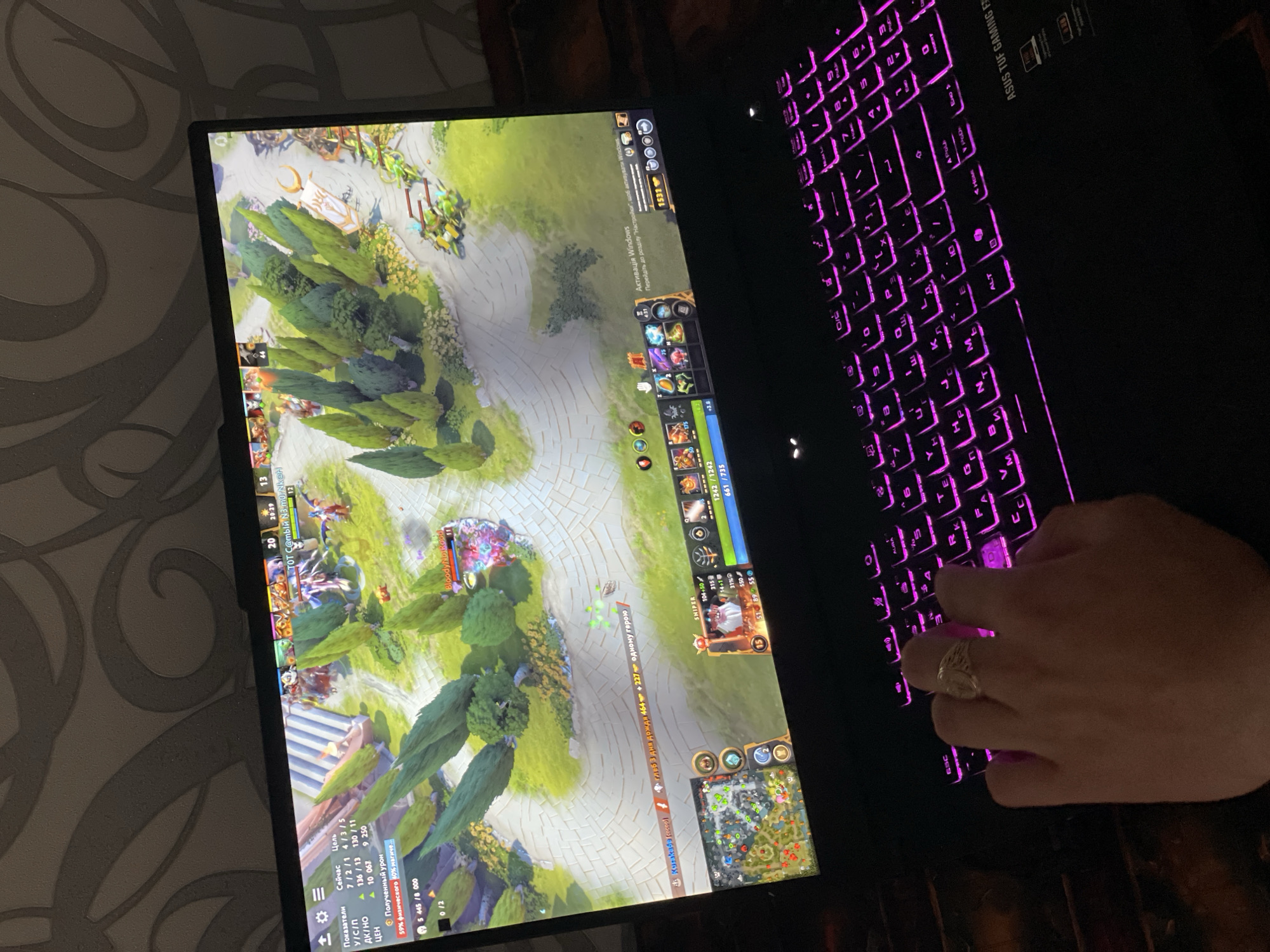 A person's hand is seen on a laptop keyboard with purple backlighting. The laptop screen displays the game Dota 2. The laptop model is ASUS TUF GAMING F. On the bottom of the screen is text written in Russian or Ukrainian. At the top of the screen is text that reads "TOT C@тый №3 m0rNk @H". There is an activation notice for Windows at the top of the screen. The image was taken indoors. The city of Rozsoshentsi, Ukraine, is not visible in the image.