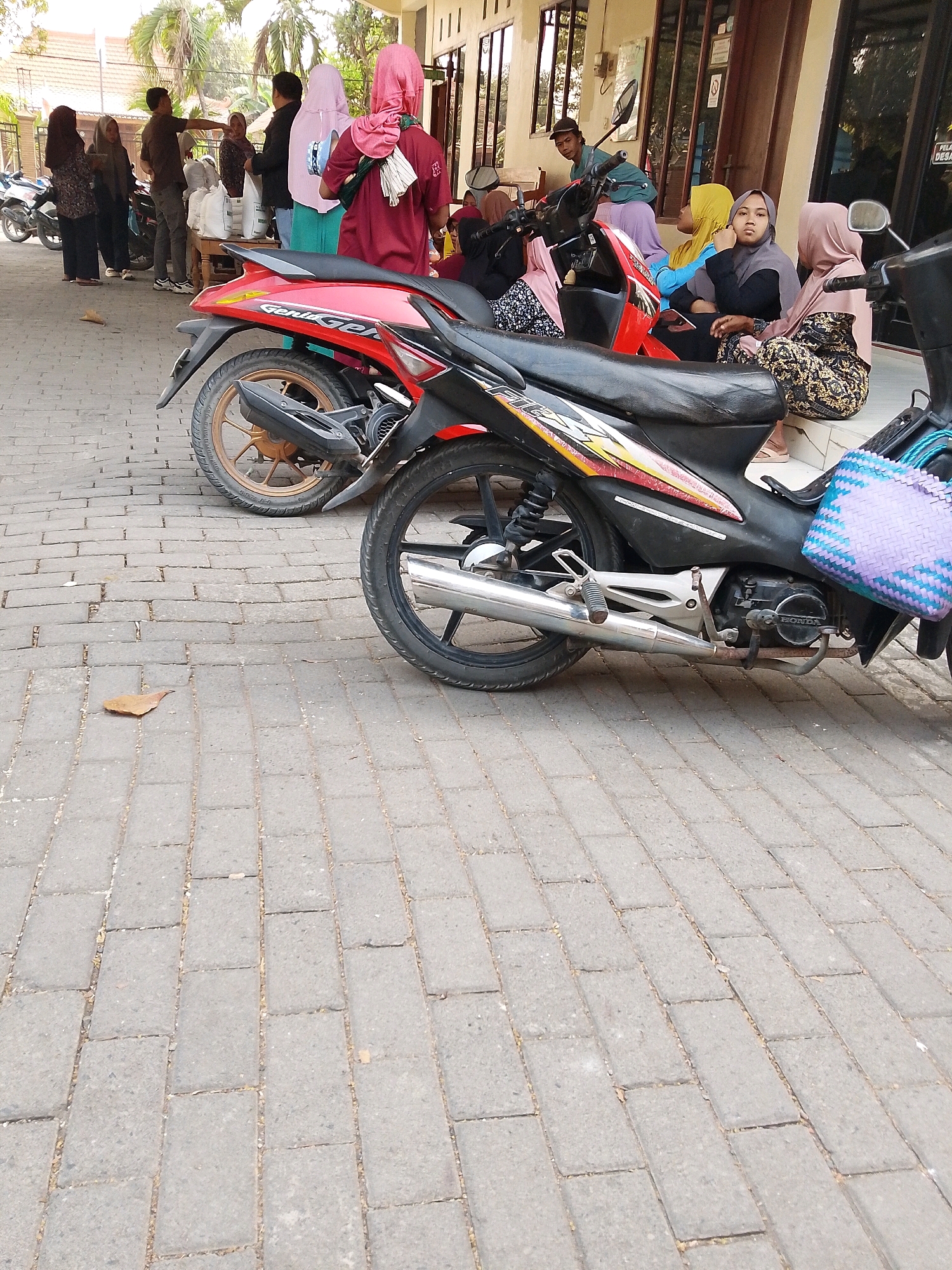 In Wadang, Indonesia, the image captures a street scene with several people standing and sitting near a building with a row of windows. Two motorcycles are prominently parked on a paved brick surface. The people are dressed in casual clothing, with several women wearing headscarves. Some individuals appear to be engaged in conversation or waiting. The environment suggests a public area, possibly near a market or community center, with a portion of the building's facade visible. The presence of motorcycles indicates this is a mode of transportation in this location.
