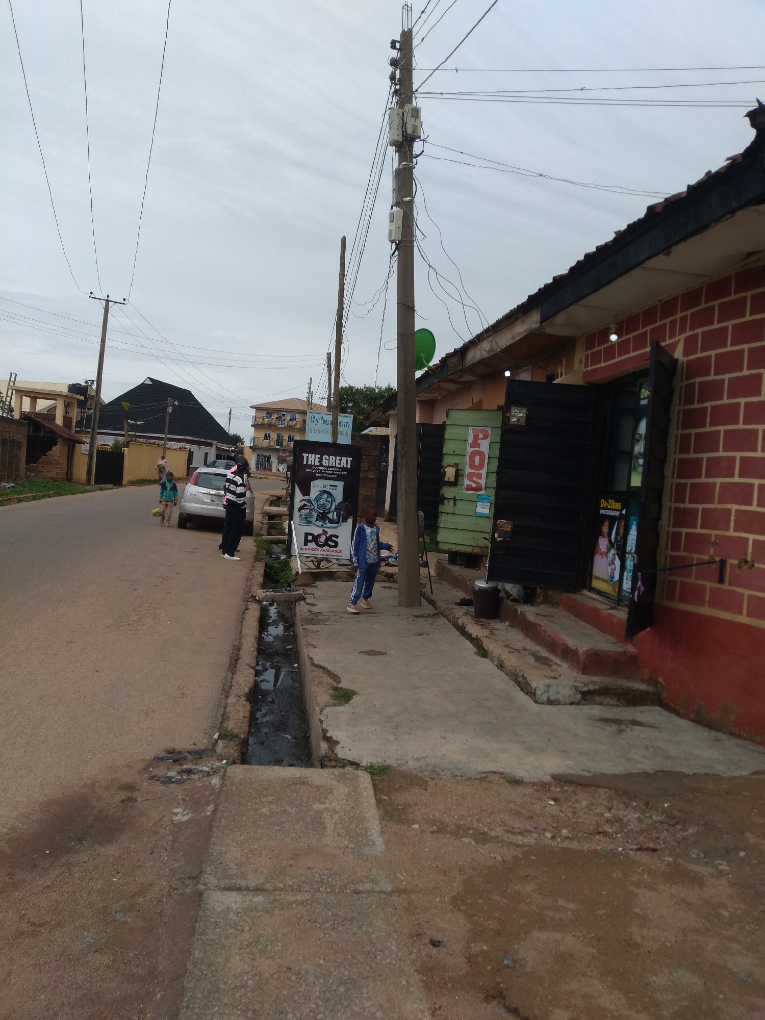 This media file depicts a street scene in Jos, Nigeria, under an overcast sky. The foreground features a dusty road with a concrete drainage ditch running alongside it. Several people are present: a man in a striped shirt and dark pants stands near a silver car, a boy in a blue tracksuit walks on the sidewalk, and two children are visible further down the road.
The background shows buildings, including a red brick structure with an open storefront, and other residential or commercial buildings. A tall utility pole with multiple wires and electrical boxes is prominently positioned. Several signs are visible, including one for "THE GREAT POS SERVICES AVAILABLE" and another for "De-Zibas PHOTOGRAPHY." The overall impression is of a typical day in a bustling urban environment, with everyday activities taking place. The weather appears to be cloudy, suggesting it might be later in the day or a generally overcast period. The presence of the drainage ditch and the dusty road hints at the climate and infrastructure of the location.