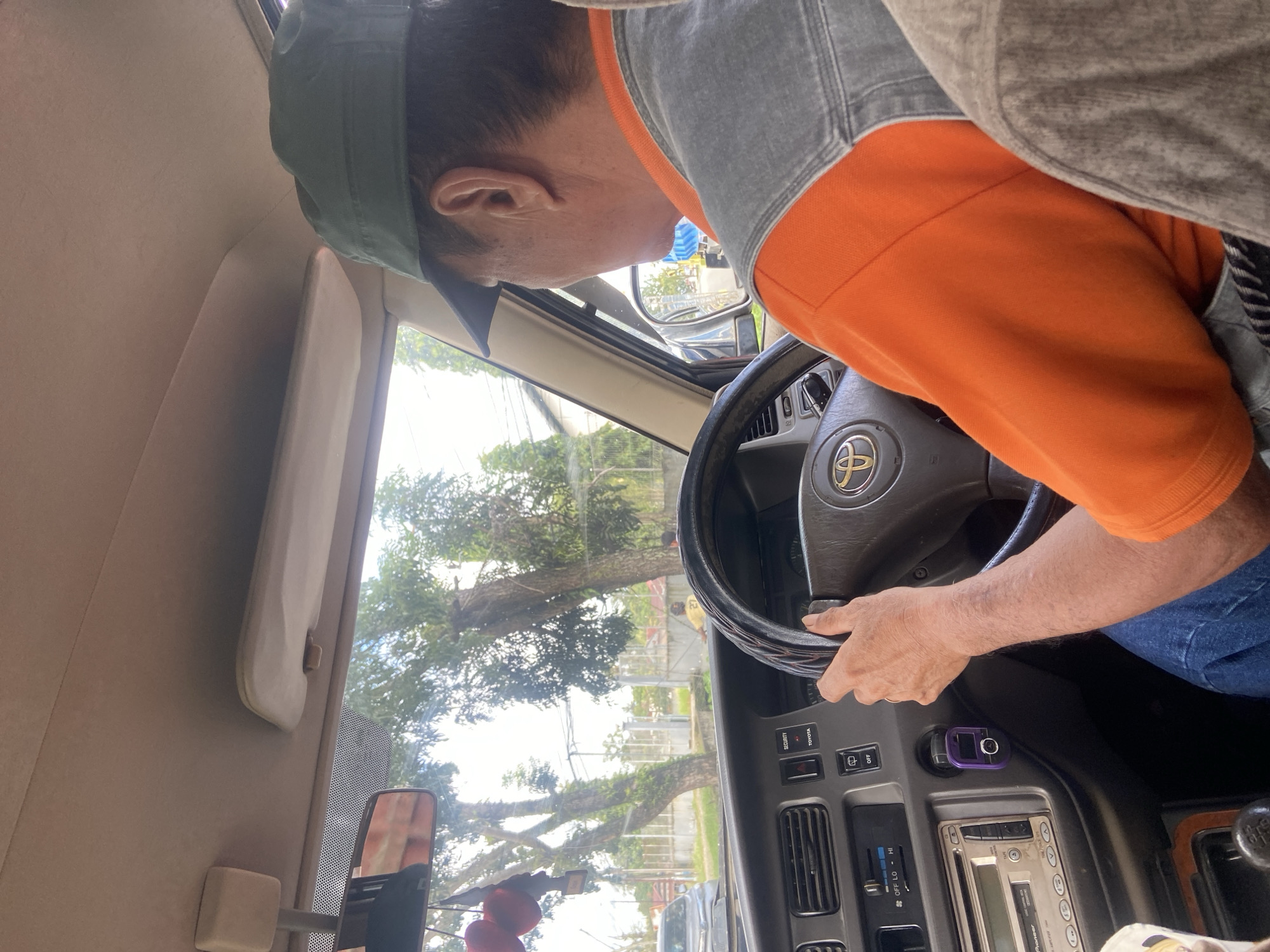 In the interior of a car in Palopo, Indonesia, a man wearing an orange shirt and a grey vest, along with a green cap, is driving. The car's dashboard is visible, featuring a Toyota steering wheel, a car radio, and various buttons and controls. Outside the car, trees and buildings can be seen through the windows, suggesting an urban or semi-urban environment. The scene appears to be captured during daytime.