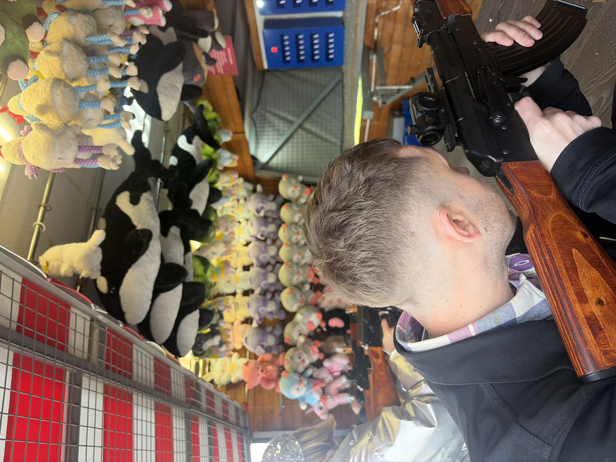 This image depicts a man participating in a shooting gallery game, likely at an amusement park or carnival in Saint Petersburg, Russia. The man, viewed from behind his head and right shoulder, is holding an airsoft or pellet rifle, which resembles an AK-47 and is fitted with a red dot sight. He appears focused, aiming the rifle.
The background reveals a vibrant array of stuffed animal prizes lining the walls of the booth. These include numerous white sheep with colorful rope-like legs, black and white cow-like plush toys, and a variety of other soft toys like pigs, bunnies, and unicorns in different colors. Above some of the prizes, blue electronic panels with indicator lights suggest a scoring or target system. The game booth features wooden structures and a red and white striped barrier at the foreground, common in fairground attractions. The lighting suggests an indoor or covered setting. Another person's hair and shoulder are partially visible at the bottom of the frame.