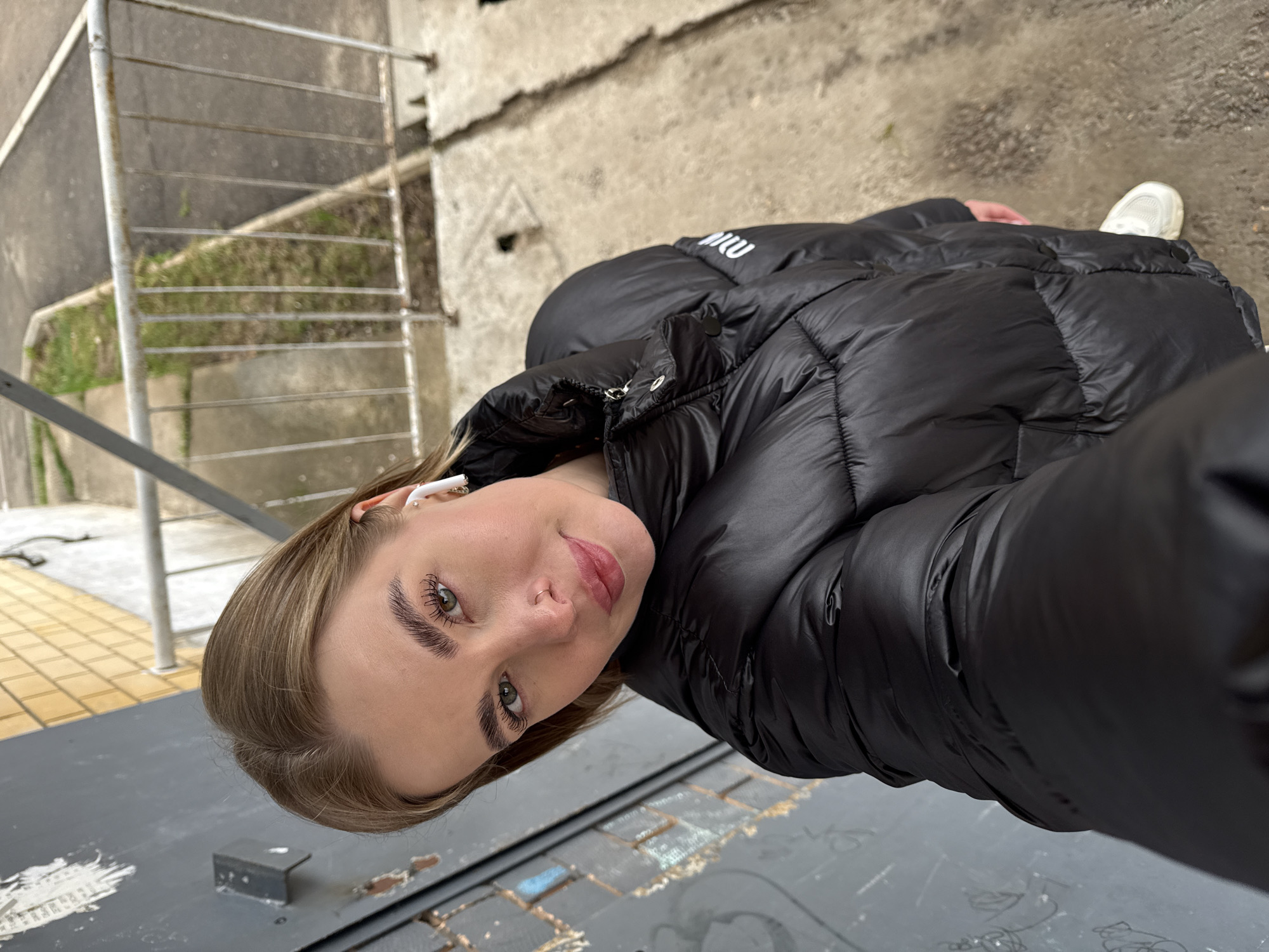 A young woman is taking a selfie outdoors. She is wearing a black puffer jacket and has white earbuds in her ears. She stands on a grey surface, likely a street or sidewalk. In the background, there is a set of stairs with a metal railing. Behind the stairs, there is a concrete wall and some greenery. The location is Chaikovsky, Russia.