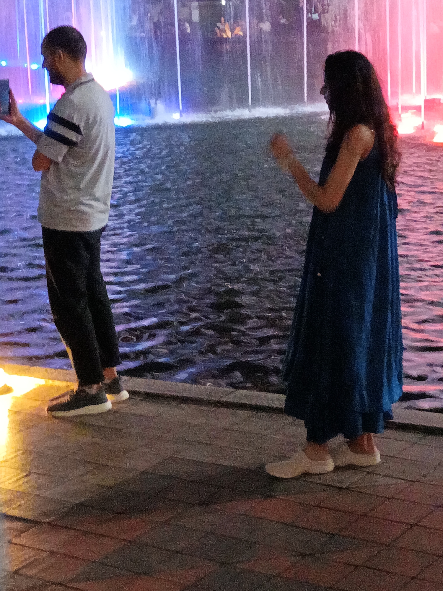 This is a medium shot, low-angle photograph of a couple standing near a water feature with colorful lights, likely taken at night in Kuala Lumpur, Malaysia. The couple is positioned on a textured, tiled walkway.
On the left, a man with dark hair and a beard is standing, facing left and slightly away from the camera. He is wearing a grey, short-sleeved polo shirt with dark stripes on the sleeves and dark pants. He is holding a rectangular object, possibly a phone, in his left hand. His right arm is bent at the elbow, with his hand resting near his waist.
To the right of the man, a woman with long, dark, curly hair is standing, facing left and slightly towards the camera. She is wearing a long, flowing, dark blue dress. Her arms are bent, and her hands are raised, with her right hand extended slightly forward.
The background features a water fountain with cascading water illuminated by vibrant colored lights, primarily in shades of blue, pink, and white. The water surface is choppy and reflects the lights. The ground beneath the couple is a paved area with interlocking rectangular tiles in shades of red, brown, and grey, with a bright yellow light casting a strong reflection on the left side of the frame. The overall impression is a contemporary urban setting at dusk or night.