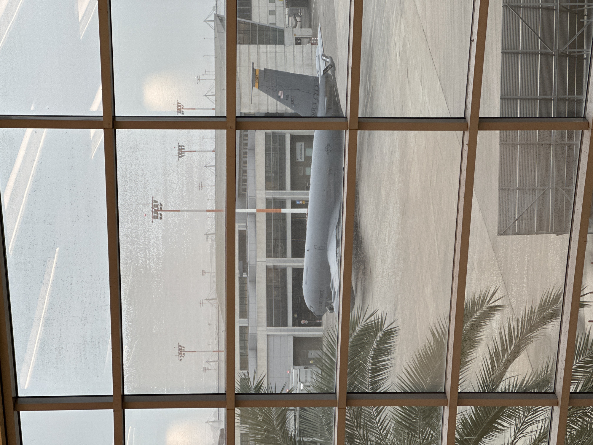 The image shows a view from inside a building, looking out through a grid of windows, likely at an airport. Visible through the windows is an aircraft, specifically a KC-135 Stratotanker of the US Air Force, discernible by its tail markings "IOWA" and the American flag, along with the tail number "22555". The aircraft sits on the tarmac, adjacent to what appears to be an airport terminal building. Parts of the airport infrastructure, such as lighting poles, are visible. The window is partially obscured by what seems to be condensation or rain, adding a slightly blurred effect to the scene. In the foreground, portions of palm tree branches are present, possibly obscuring the lower part of the view. Given the geographical location of the KC-135 Stratotanker, this may not be in Tel Aviv-Yafo, Israel, as US Air Force tankers are not commonly stationed there.