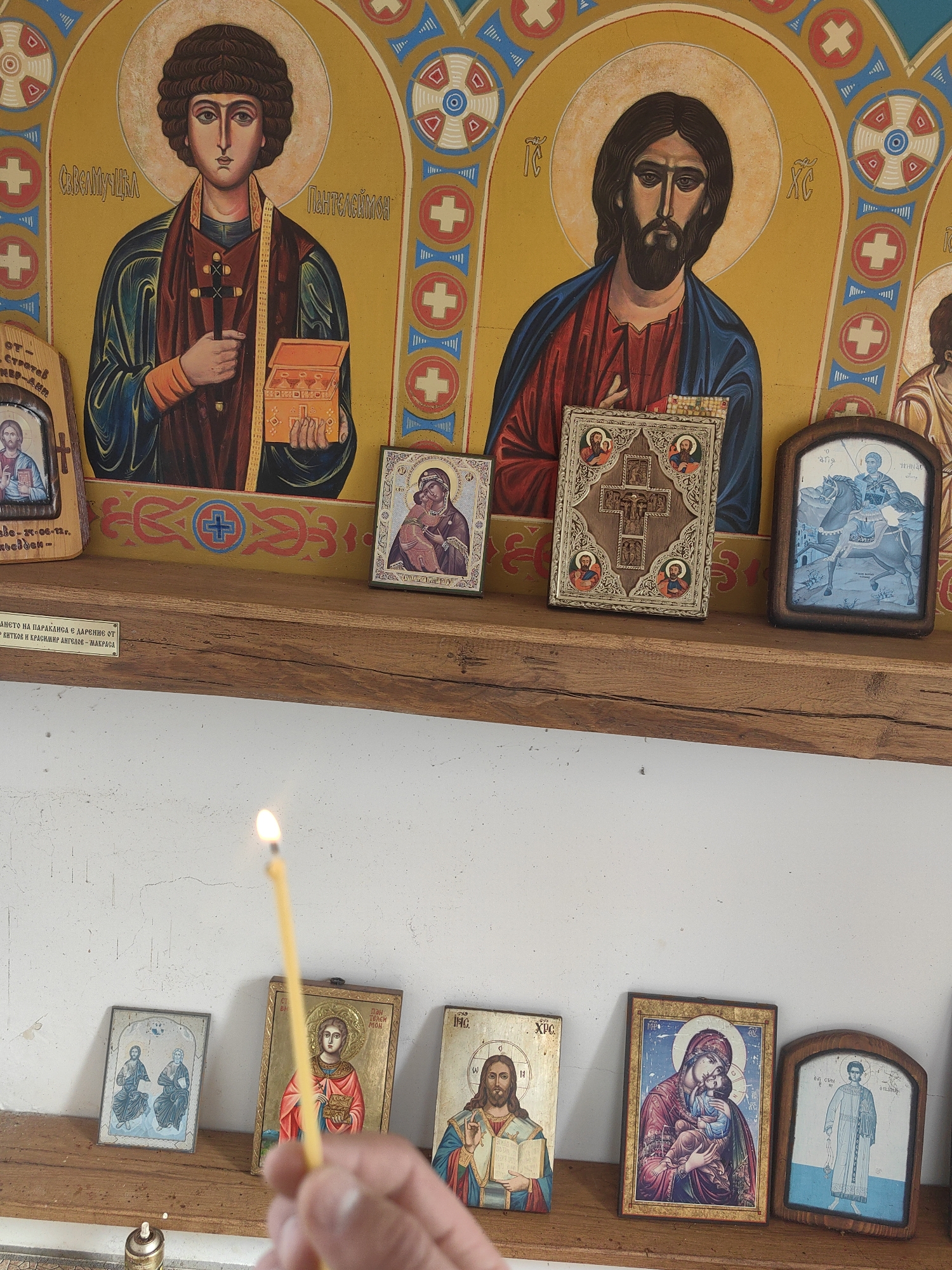 The image depicts an assortment of religious icons and candles on wooden shelves, likely in a church or similar religious setting. A hand holding a lit yellow candle is visible in the foreground. The upper shelf displays larger icons with religious figures, while the lower shelf holds smaller icons of similar subjects. There are labels or inscriptions in Cyrillic on some of the items, suggesting a Bulgarian context. Based on the OCR, the image appears to be from Gumoshtnik, Bulgaria.