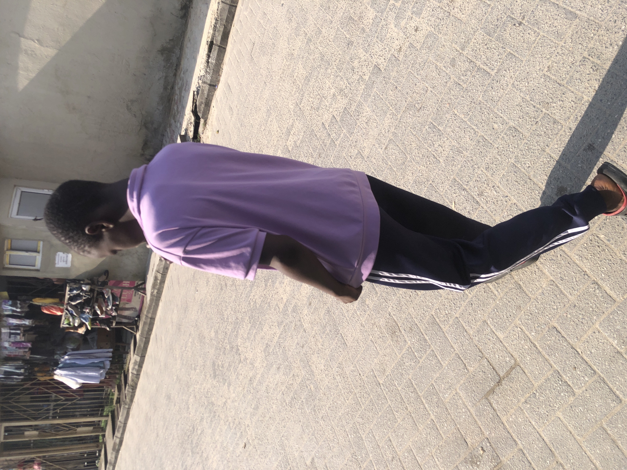 A young male, viewed from the back, stands on a paved sidewalk in Ado, Nigeria. He is wearing a lavender polo shirt and dark blue track pants with white stripes. He appears to be walking or dancing, with his right arm slightly bent at the elbow. Behind him, a row of shops is visible, featuring clothing racks with shirts and a shoe display stand. A light-colored wall forms the background on the right side of the image. The sun casts a shadow from the individual onto the pavement.