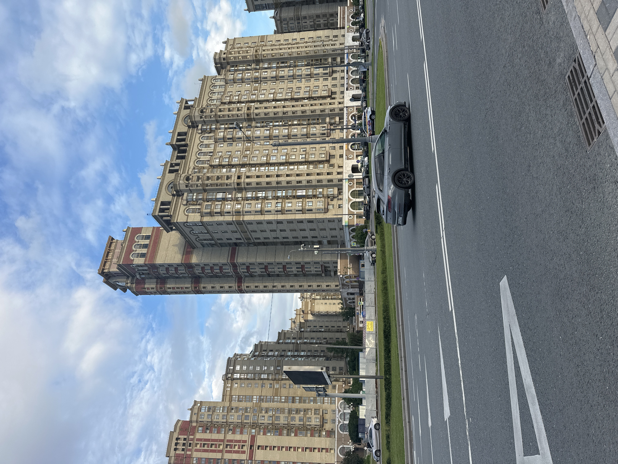 The image depicts a street scene in Moscow, Russia, featuring modern residential buildings and a multi-lane road. The buildings are tall, constructed with a combination of light-colored facade and red-brick accents, exhibiting a uniform architectural style with numerous windows and decorative elements. In the foreground, a grey station wagon is driving on the asphalt road, which is marked with white lane lines and arrows. A grassy median separates the road from a sidewalk area in front of the buildings, where several cars are parked. The sky is partly cloudy, with a mix of blue sky and white clouds, suggesting daytime. The overall environment appears to be an urban setting, likely a residential or mixed-use district.