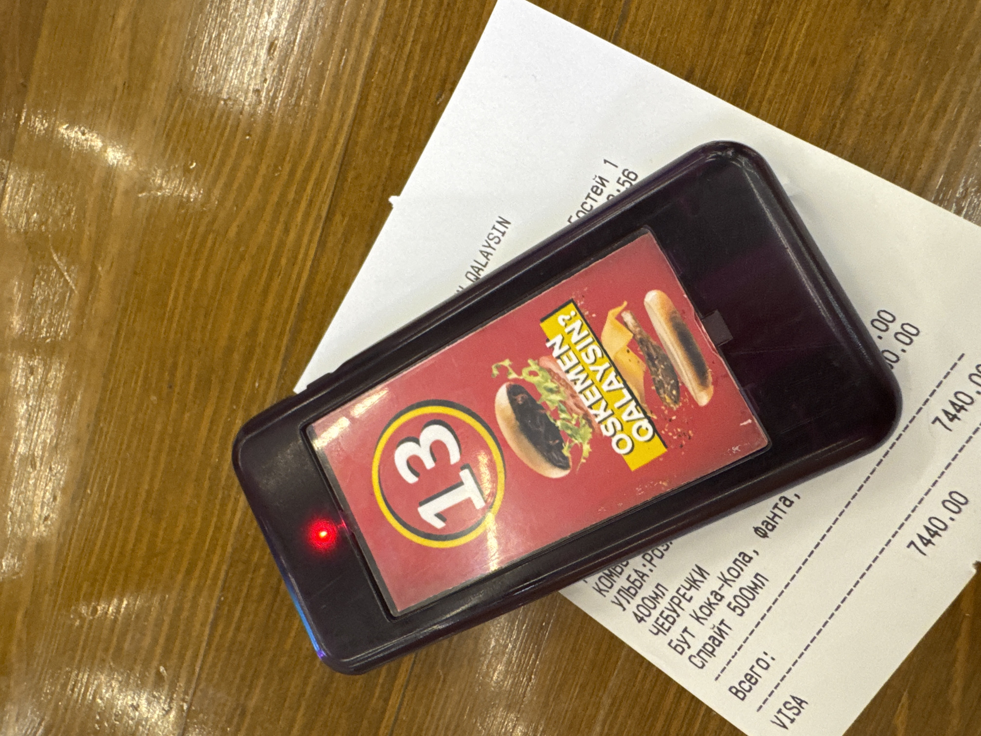 An overhead medium shot shows a dark purple order pager with a red indicator light on top, placed on a white receipt on a polished wooden table. The pager screen displays a large yellow-circled number "13" in white, superimposed on a red background with an image of a burger. Text below the burger image reads "OSKEMEN QALAYSIN?". The receipt features handwritten and printed text, including "VISA", a total of "7440.00", and other items that appear to be food orders such as "КОМБ", "УЛЬБА", "400мл", "ЧЕБУРЕЧКИ", "БУТ Кока-Кола, фанта", and "Спрайт 500мл". The location is identified as Oskemen, Kazakhstan, based on the text on the pager. The scene is static, with no people or actions visible.