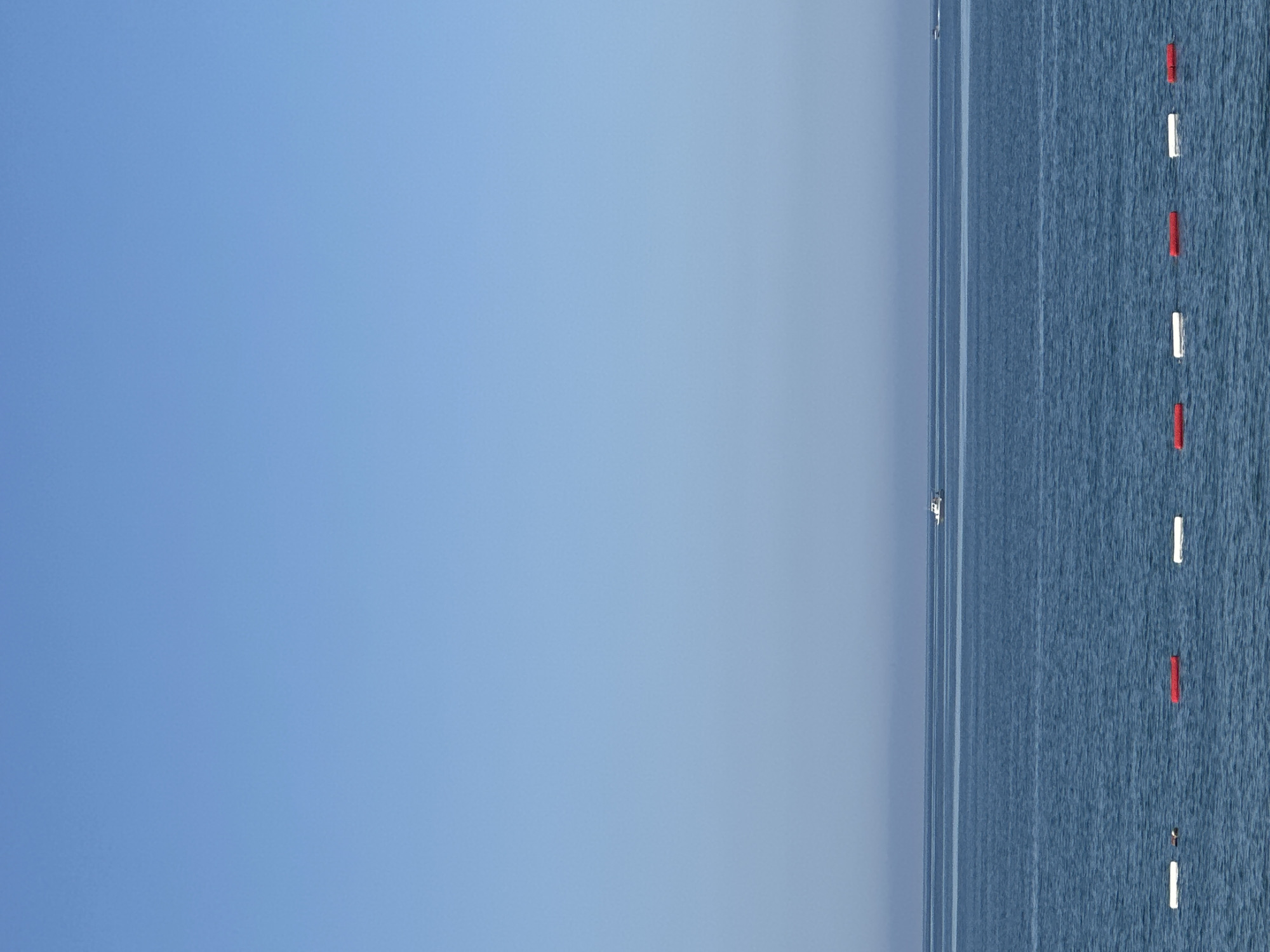 The image shows a view of the Adriatic Sea near Bar, Montenegro. The sky is a clear, gradient blue. To the right, the sea displays a textured surface, marked by small waves. A line of alternating red and white buoys floats in the water, indicating a boundary. Further out, a small boat is visible on the horizon.