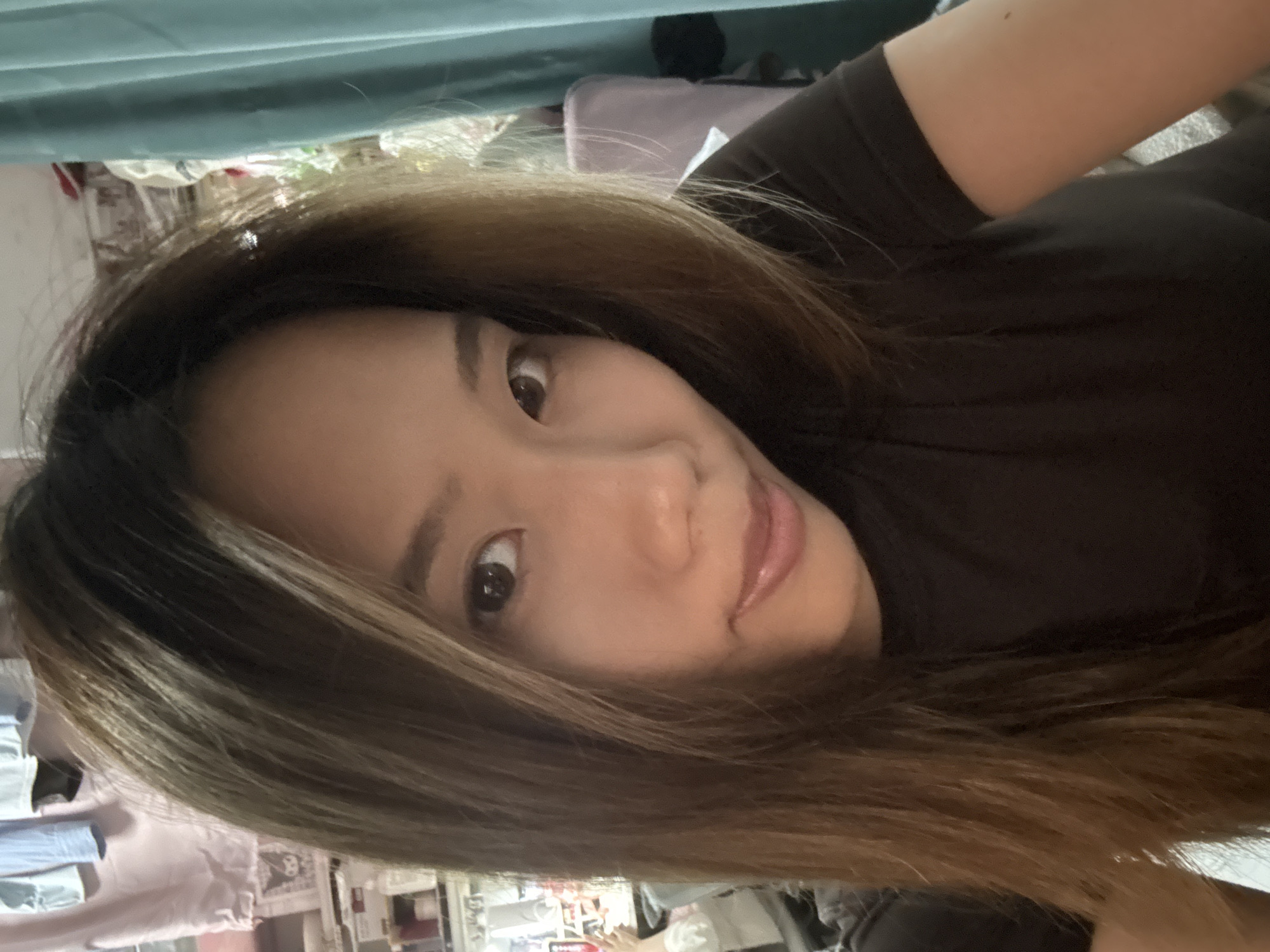A woman with long brown hair and light skin is looking directly at the camera with her head slightly tilted. She is wearing a dark brown shirt. The background is cluttered and out of focus, with a mix of objects and materials including clothing, bedding, and furniture. The location is in the Nan'an District in China. The precise location within the city cannot be determined from the image.