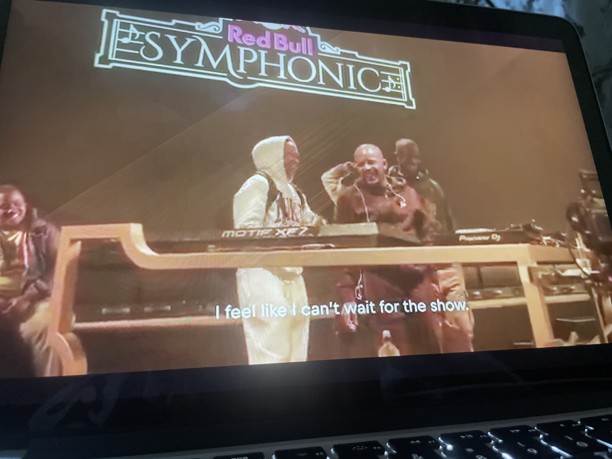 The image shows a laptop screen displaying a video of a live event. At the top of the screen, a logo reads "Red Bull Symphonic". Below the logo, several individuals are visible on a stage, appearing to be performers or DJs. In the foreground, a person wearing a white hooded jacket is standing behind a console with what appears to be DJ equipment. To their right, another person, shirtless and wearing headphones, is also interacting with the equipment. Further back, other individuals are present. At the bottom of the screen, white subtitles read, "I feel like I can't wait for the show." The overall environment appears to be a concert or performance venue. The image is taken from a perspective looking down at the laptop. There is no visible information in the image that provides a specific location in the city of Howayi, Nigeria.