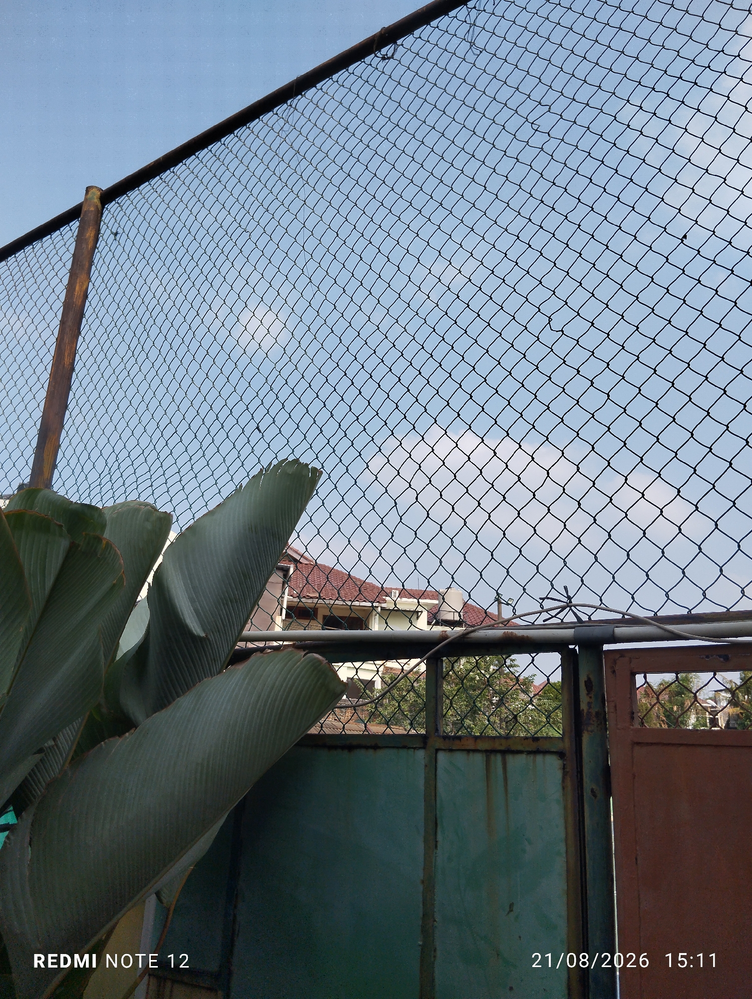 The image captures an outdoor scene in Bekasi, Indonesia, featuring a chain-link fence with a weathered brown metal gate to the right. Behind the fence, a building with a red roof is partially visible. In the foreground, large green leaves from a tropical plant are prominently displayed, obscuring part of the fence and gate. The sky above is a pale blue with scattered clouds. A timestamp in the lower right corner reads "21/08/2026 15:11," and "REDMI NOTE 12" is textually watermarked in the lower left. The overall impression is one of a slightly overgrown urban or suburban environment.