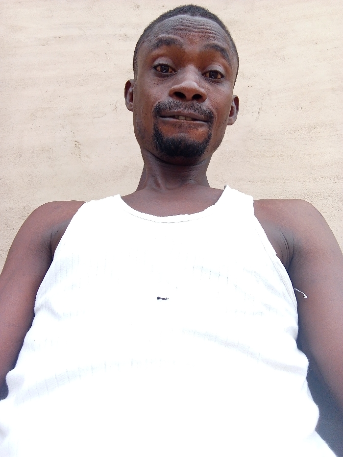 A low-angle, close-up photograph captures an adult male of African descent, positioned against a plain, light beige textured wall. The male subject has short dark hair and a dark goatee beard. He is wearing a white ribbed tank top, on which a small, dark, horizontally elongated mark is visible on the upper left chest area. His eyes are dark, and he looks directly at the camera with a neutral expression. The scene is located in Adonkolo, Nigeria.