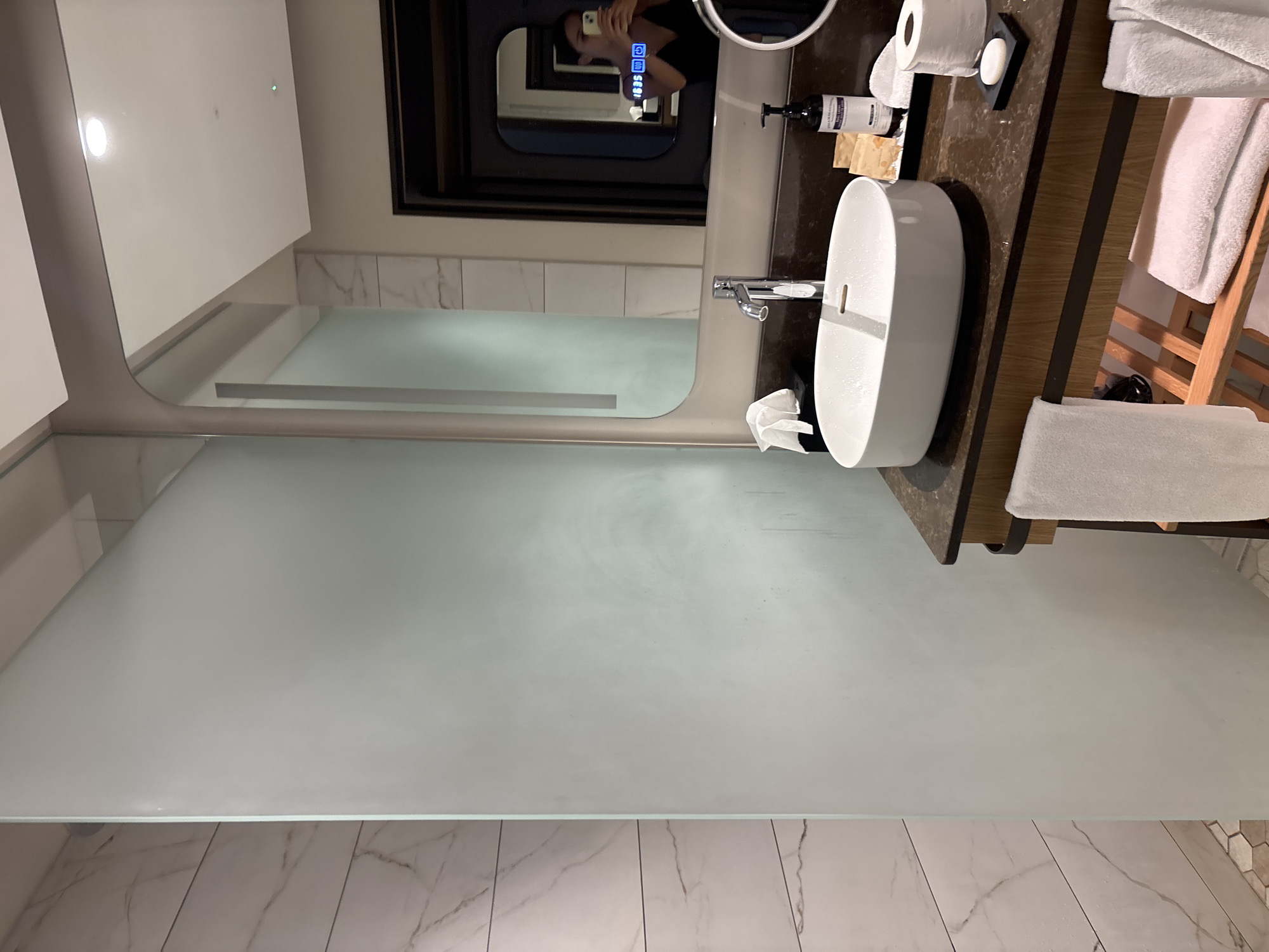 The image shows a modern hotel bathroom with marble tiling and a frosted glass partition. In the foreground, a white oval sink with a chrome faucet sits on a dark countertop. To the right of the sink are a bottle of soap, individually wrapped amenities, a roll of toilet paper, and a bar of soap on a black dish. A white towel is draped over the vanity. Below the vanity, shelves hold folded white towels. The background features a large mirror reflecting a portion of the room and a person taking a selfie. The scene is set in a bathroom in Razliv, Bulgaria.