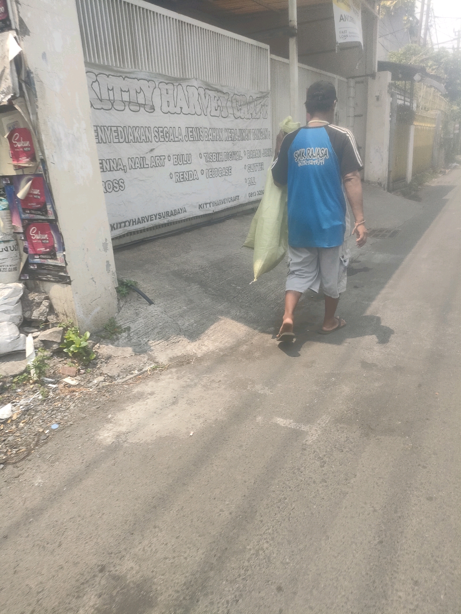 A man wearing a blue and black t-shirt with the words "SMK RAJASA SURABAYA" printed on the back and gray shorts walks away from the camera on a paved road. He is carrying a large, light-colored sack over his right shoulder. To his left is a white wall with a banner that reads "KITTY HARVEY CRAFT" and lists various items such as "ENNYEDIKAN SEGALA JENIS BAHAN KERAKYATAN", "ENNA, NAIL ART, BULU, TASBIH DIGITS, DLL", and "ROSS, RENDA, DECOCASE". The banner also includes the text "KITTYHARVEYSURABAYA" and a phone number "0813 3292". There are some white sacks and rocks to the left of the banner, and some vegetation growing near the base of a pillar. The environment appears to be a street in Surabaya, Indonesia, with a building and fence visible in the background. The overall lighting suggests it is daytime, possibly in the morning or afternoon.