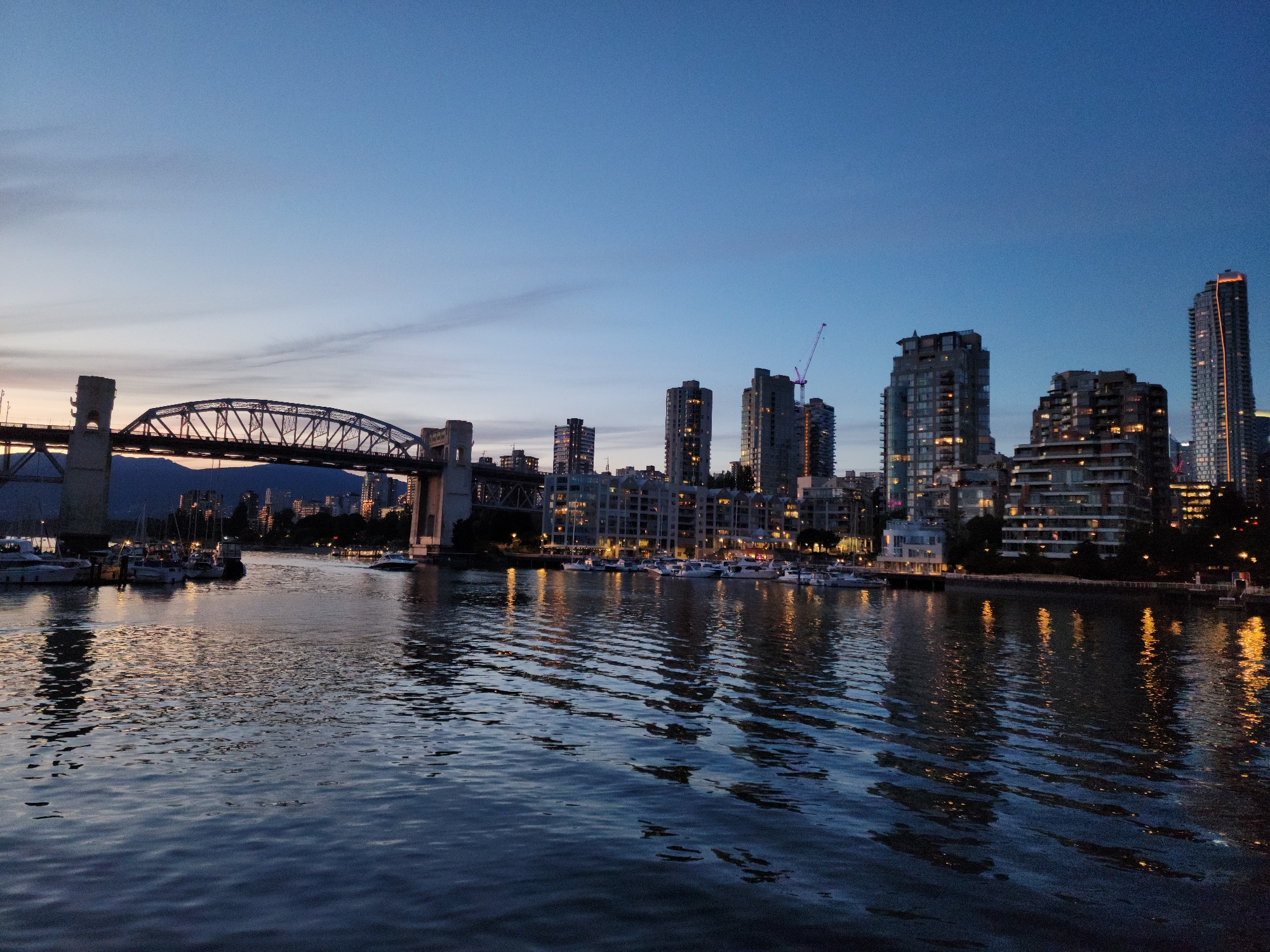 The image depicts a city skyline at dusk, likely Vancouver, British Columbia, Canada. A large steel arch bridge spans the water in the left foreground, with its towers illuminated by artificial lights. Several sailboats and yachts are docked on the water near the bridge. The background features a cluster of high-rise buildings, some of which are illuminated with warm interior lights, and a construction crane is visible on top of one of the buildings. The water in the foreground reflects the lights from the city and the sky, creating a shimmering effect. The sky is a deep blue, transitioning to a lighter hue near the horizon where the last vestiges of daylight are visible. The overall mood of the image is serene and tranquil, capturing the beauty of the city as day turns into night.