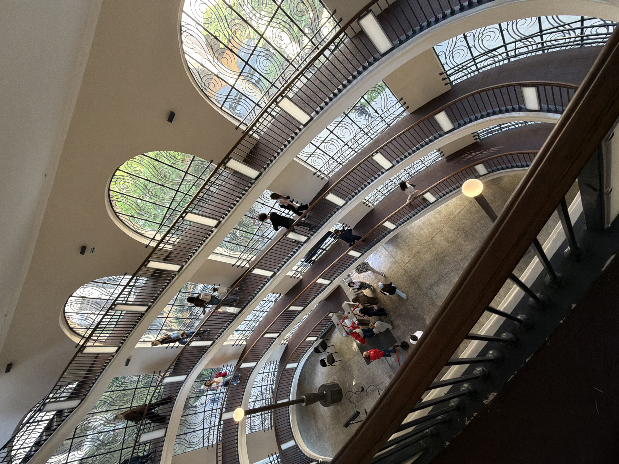 The image presents a high-angle view looking down into the interior of a multi-story building, featuring a grand, sweeping, curved staircase. The architecture is characterized by its elegant, flowing lines and multiple levels.
The central feature is a wide, carpeted staircase with dark metal railings topped with a polished wooden handrail, spiraling down towards the ground floor. Numerous large, arched or circular windows punctuate the walls of the building, each adorned with intricate, scrolled metal grilles that cast decorative shadows. Through these windows, bright daylight and lush green foliage are visible, indicating the time of day is daytime.
Several people are scattered across different levels. Individuals are seen walking or standing on the stair landings and steps. On the spacious ground floor, a larger group of people is gathered. Some are standing, while others are seated on chairs or appear to be lying down on mats or low platforms. A prominent dark, cylindrical column or art installation stands on the ground floor, surrounded by various pieces of equipment.
The overall scene depicts a calm and open public space, possibly a library, museum, or an historic department store, given the architectural grandeur and ornate details, particularly the window grilles. The lighting is a blend of ample natural light from the windows and artificial light fixtures, including a spherical lamp on a pole near the ground floor. No discernible text is visible within the image. The location is Tbilisi, Georgia.