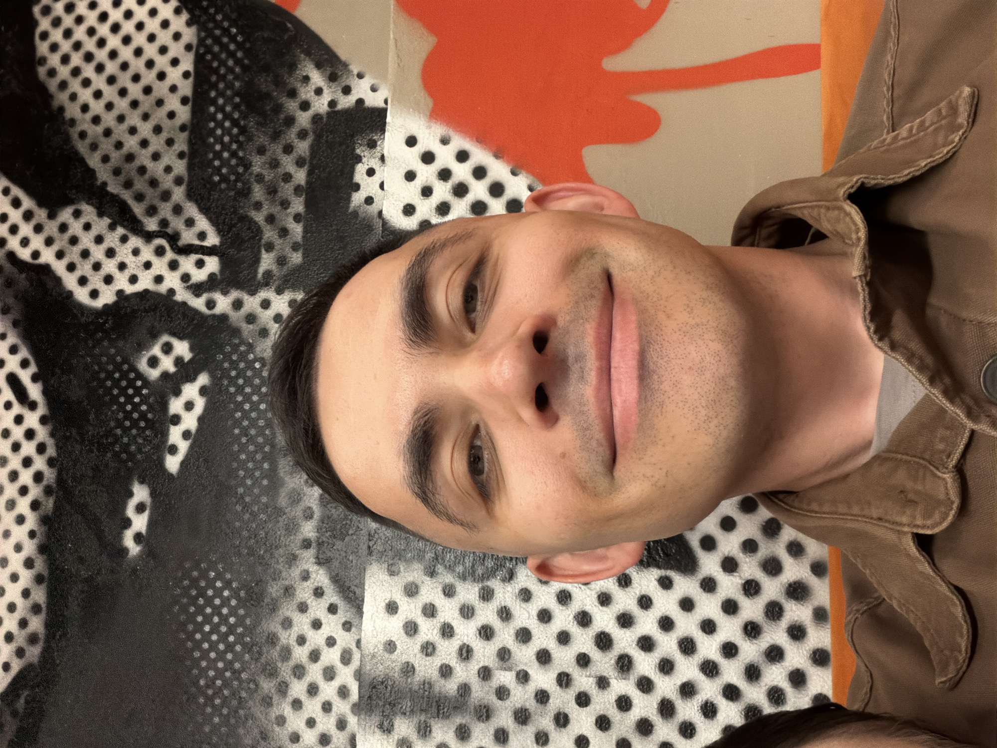 A man is looking at the camera. He has short, dark hair, brown eyes, and light skin. He is wearing a brown jacket. The background behind him is an abstract design with black and white polka dots, black and white airbrushing, and orange paint. There is no indication of the location being Izhevsk, Russia.