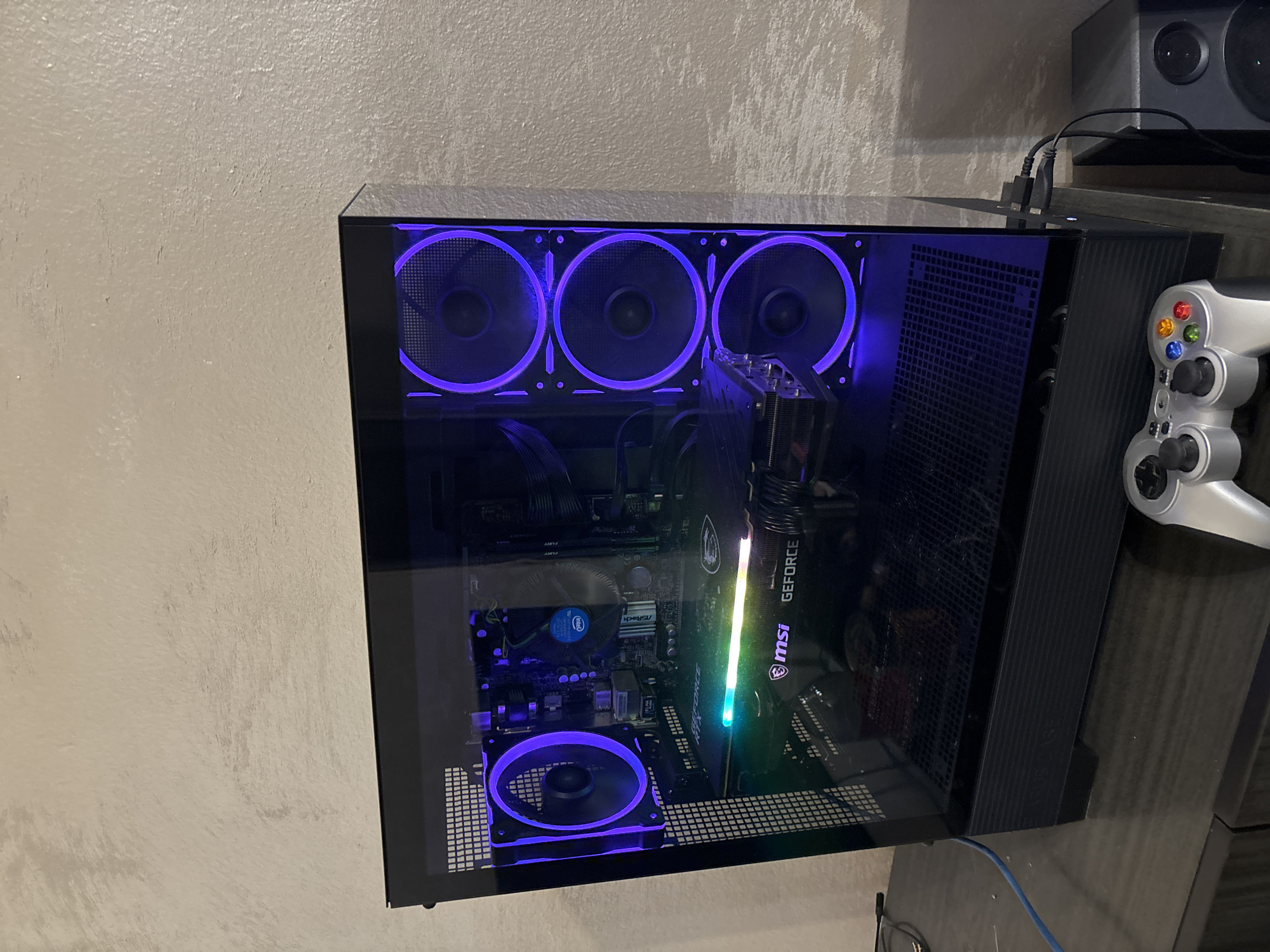 The image depicts a custom-built personal computer tower with a transparent side panel, revealing its internal components. The tower is black with blue LED-illuminated fans. Three fans are positioned at the top, and one is visible at the bottom. Key components visible inside include a motherboard with an Intel CPU, an MSI GeForce RTX graphics card, and a CPU cooler. The computer is placed on a dark-colored surface. A gray Xbox controller and a speaker are partially visible to the right of the tower. The background shows a light-colored wall. The image was not taken in Plovdiv, Bulgaria.