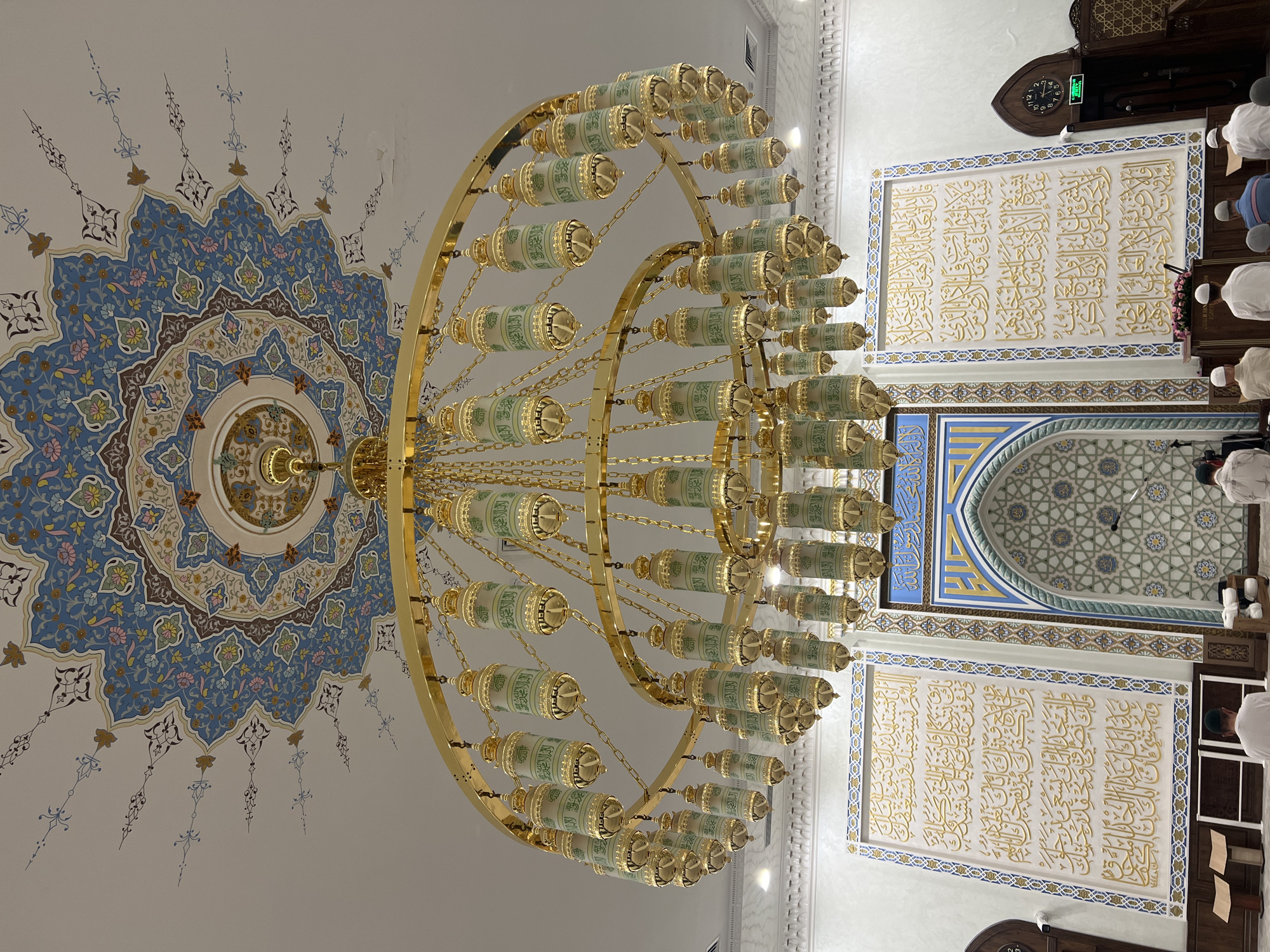 The image depicts the interior of a mosque, likely in Tashkent, Uzbekistan, characterized by Islamic architectural and decorative elements. A large, ornate gold chandelier with multiple tiers and numerous lantern-like light fixtures hangs from the ceiling. The ceiling itself is adorned with intricate, colorful Islamic geometric patterns and floral motifs in blue, beige, brown, and pink hues.
Below the chandelier, the scene shows a prayer area. A mihrab, an arched niche indicating the direction of Mecca, is visible, decorated with geometric patterns in blue, white, and gold. Flanking the mihrab are large panels featuring elaborate Arabic calligraphy in gold on a white background.
Several individuals, appearing to be men, are present in the prayer area, dressed in traditional attire, with most wearing white. Some are seen kneeling in prayer, while others are standing or seated. The overall environment suggests a place of worship during prayer time. The lighting appears to be a mix of ambient light and the illumination from the chandelier.