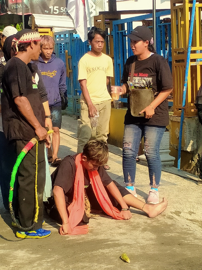 The image depicts an outdoor scene in Sepat, Indonesia, on a concrete or asphalt surface under bright sunlight. Six individuals are present. In the immediate foreground, a male wearing a black shirt, dark pants, and blue and yellow shoes, with a red and white patterned headband, stands holding a multi-colored (red, green, yellow, black) braided rope or whip in his left hand. Adjacent to him, a male with light-colored hair, dressed in a black shirt and an orange-red scarf, is seated directly on the ground, manipulating the scarf with both hands. His bare feet are visible, and a small, light-green, pointed object rests on the ground near them.
Behind the seated individual, a male in a light yellow "ZEROCAM" t-shirt and light-colored pants stands, holding a clear plastic bottle in his left hand. To his right, a female wearing a black baseball cap, a black t-shirt with a graphic design, ripped blue jeans, and light-colored athletic shoes with pink and blue socks stands, holding a clear plastic cup containing a light-colored liquid in her right hand and a dark shoulder bag in her left. Further in the mid-ground, to the left of the male with the headband, a male with light-colored hair wears a blue "Greenlight 1996" hooded sweatshirt and dark shorts. In the far background, a partially visible individual with a white cap is present. The background features blue and yellow metal structures, a white flag, and a sign displaying "PRIMA" and "RP 15.50."