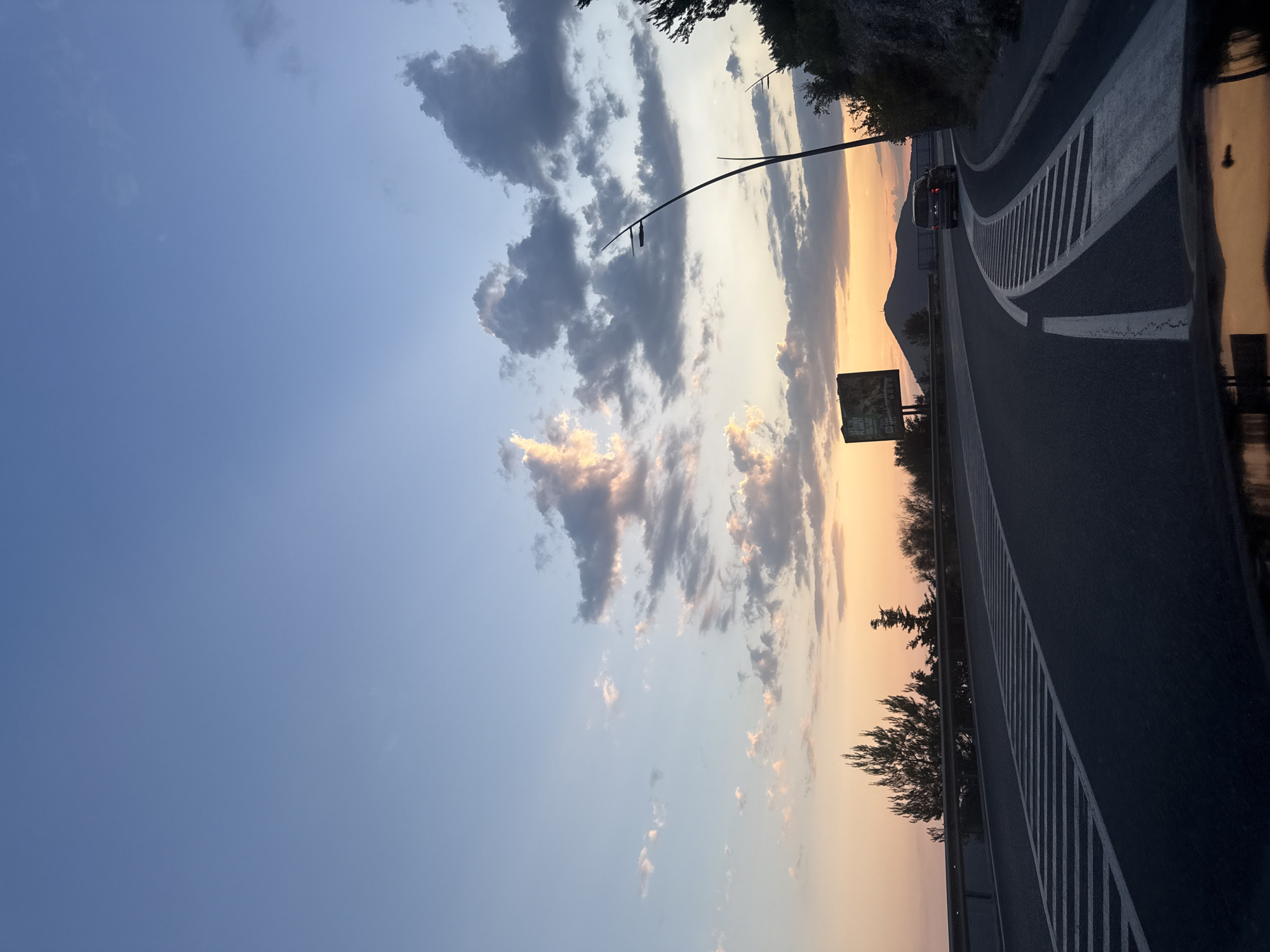 The image shows a highway at sunset, viewed from a car. The sky is a mix of blue and orange, with scattered clouds. The highway is dark and wet, with white lines marking the lanes. A car is visible in the distance, driving on the highway. Trees and a mountain are also visible in the background. The overall mood is peaceful and serene. The time of day is sunset. The weather appears to be clear with some clouds. The location is likely Kamenovo, Montenegro, given the reference to a highway and mountainous terrain. There is no visible text in the image.