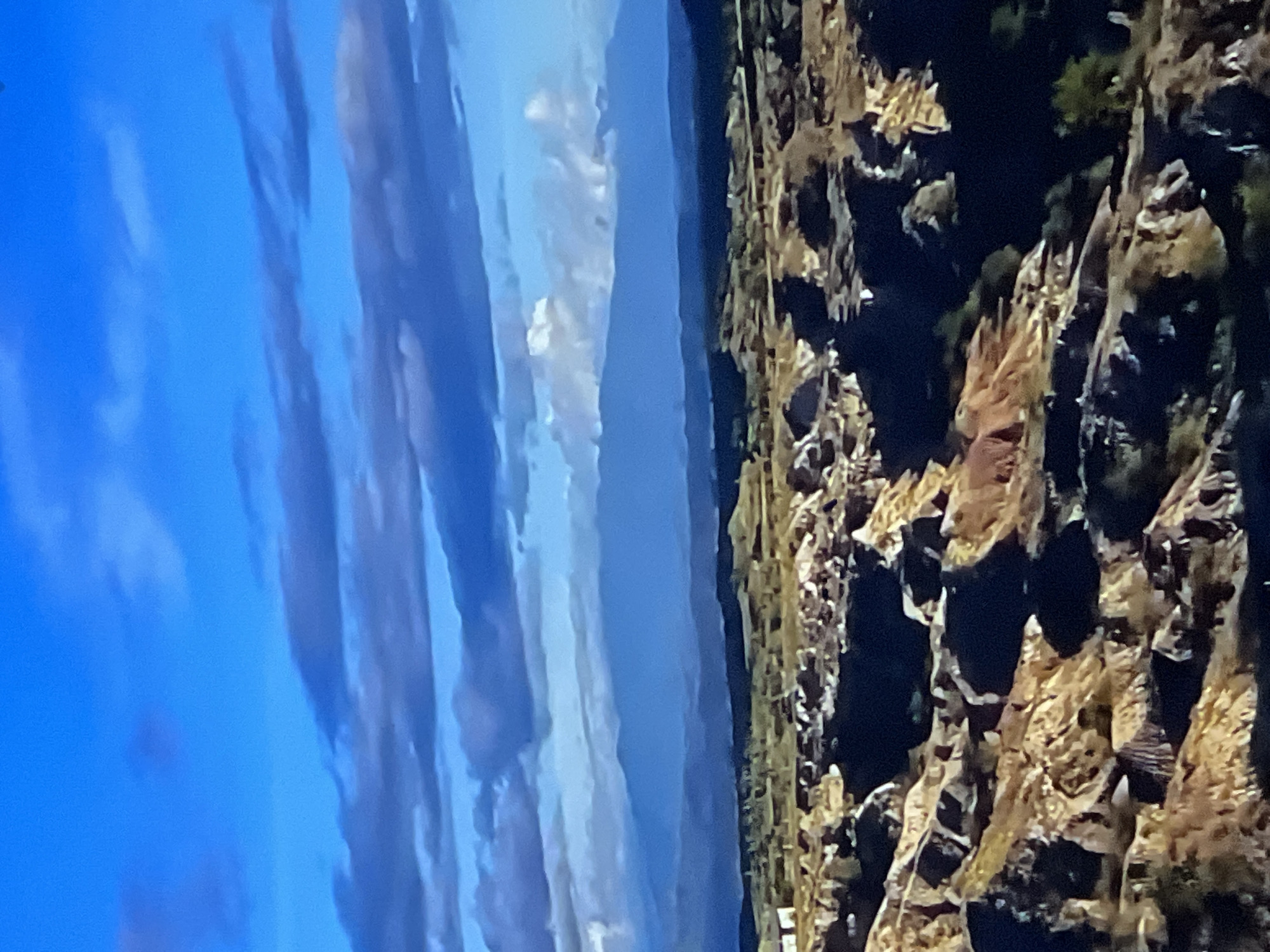 The image depicts a vast, arid landscape under a bright blue sky with scattered clouds. The foreground is characterized by rugged, rocky terrain with formations of various sizes and textures, appearing dry and eroded. Some areas are in shadow, creating stark contrasts with the sunlit portions. In the middle ground, the land flattens slightly and appears to be covered in sparse vegetation and possibly some low hills. Further in the distance, a range of mountains is visible, appearing hazy and blue in the atmospheric perspective. A faint line of darker terrain runs along the horizon before the mountains. There are no people or animals visible in the image. The scene appears to be a natural environment, possibly a desert or badlands area. The provided image does not contain any elements that would suggest it is located in Casablanca, Morocco.