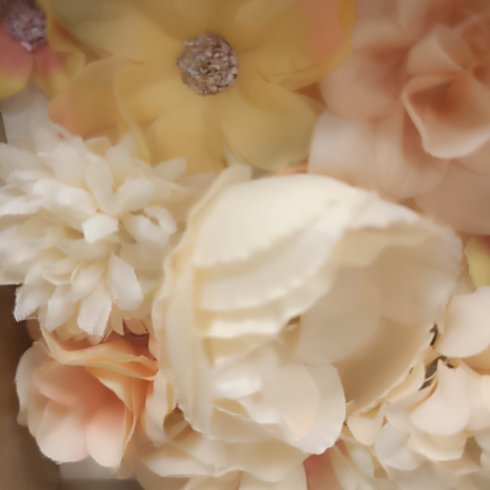The image shows a close-up of artificial flowers in various shades of cream, peach, and yellow. The flowers appear to be made of fabric and have a soft, textured appearance. Some of the flowers have decorative centers with a glittery or beaded material. The arrangement is densely packed, with overlapping petals and leaves creating a lush and full visual effect. The background is softly blurred, suggesting a shallow depth of field, which keeps the focus on the floral details. There are no people or discernible actions depicted in the image. The overall impression is one of a decorative floral arrangement, possibly intended for an event or as a home decor item. The image does not provide any information about its location in Offenbach am Main, Germany, or any other geographical context.