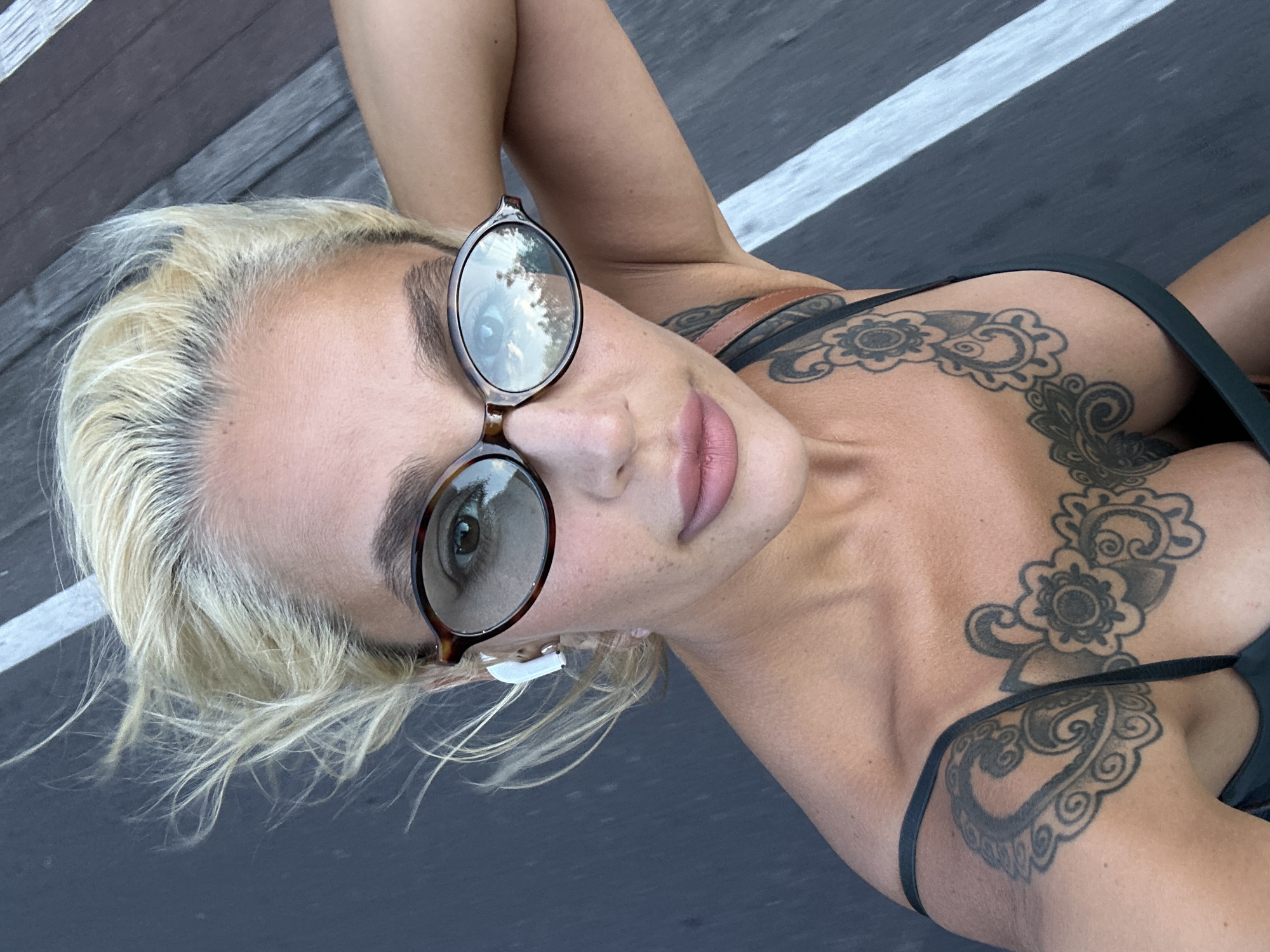 The image captures a close-up, angled portrait of a woman in an outdoor setting. She has bleached blonde hair with darker roots, dark eyebrows, and full lips with light pink lipstick. She wears tortoiseshell-framed sunglasses, which reflect trees and sky, confirming an outdoor environment. A small white earbud is visible in her left ear. A prominent black ink tattoo, featuring a floral and paisley pattern, covers a significant portion of her right shoulder and chest, partially obscured by a dark-colored strap of her attire.
The woman is positioned with her head tilted, looking directly towards the camera. The background in the upper left shows wooden planks, possibly part of a deck or boardwalk. The rest of the background is a dark grey or black paved surface with a distinct white stripe running diagonally, suggesting a road, path, or sports court. The scene appears to be taking place during the daytime, given the use of sunglasses and the reflections. The overall context suggests a casual moment in a warm climate, consistent with the hint of Kuta Selatan, Indonesia. No visible text is present in the image.