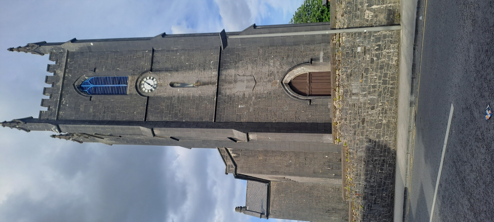 An exterior, low-angle, vertical, full shot captures the imposing stone tower of St. Patrick's Church in Castlebar, Ireland, under a partly cloudy sky. The tower, constructed of dark, weathered stone blocks, features a distinctive arched blue window near its apex. Below the window, a clock face with black Roman numerals on a white background is visible. A small, narrow niche containing a figure is situated below the clock. A smaller, attached section of the church, also made of stone, is visible to the left, with a dark slate roof. The tower and church are bordered by a low stone wall, with sparse green vegetation growing along its top. In front of the wall is a grey paved sidewalk, and further forward, a tarmac road with yellow and white markings. A lamppost stands to the right of the tower. A piece of litter, possibly a candy wrapper, is visible on the road surface in the foreground. No people are present in the scene.