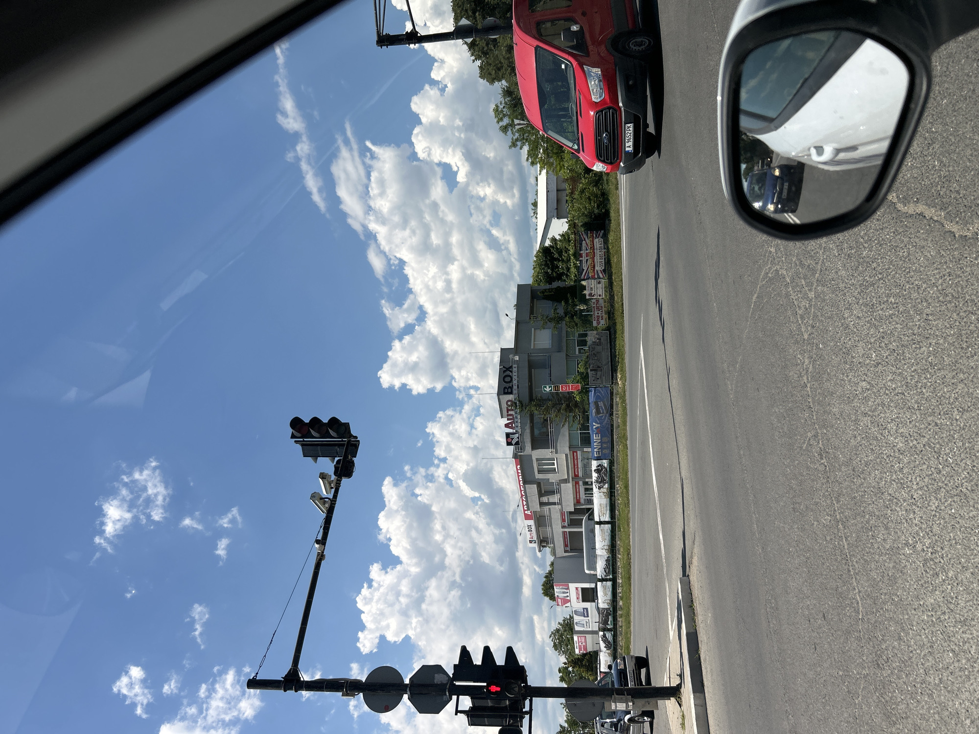 The image captures a view from a vehicle on a sunny day, looking out towards a city street and buildings. A traffic light with a red signal is visible on the left, accompanied by two surveillance cameras mounted on the traffic light pole. In the middle ground, a grey building with the "AUTO BOX" logo is prominent, along with other businesses. A red van is driving on the right side of the road, displaying a license plate reading "A 9652 PK". The sky is blue with scattered white clouds. The scene is set in the city of Nesebar, Bulgaria, near the "Auto Box" business. The image is taken from a side mirror perspective, reflecting a blue car behind the viewer's vehicle.