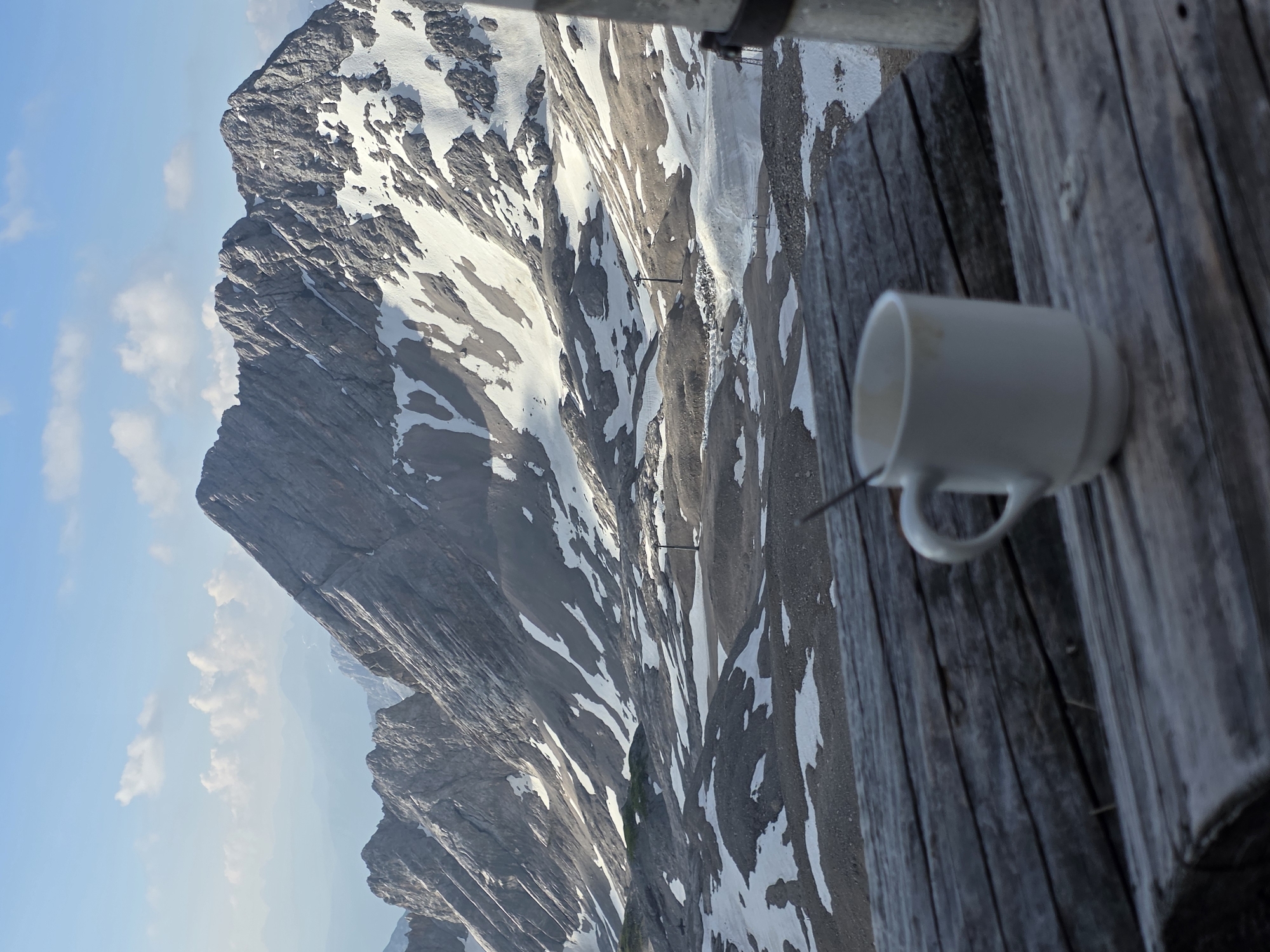 A close-up shot focuses on a white mug with a spoon resting on a rustic wooden surface. The mug is positioned in the foreground, slightly blurred, with the handle clearly visible. Behind the mug, a dramatic mountain landscape unfolds under a clear blue sky dotted with a few fluffy white clouds.
The mountains are rugged and imposing, with steep, rocky slopes partially covered in patches of snow. The snow appears somewhat melted, suggesting it's not the peak of winter. The lighting indicates it is likely daytime, possibly late afternoon, given the soft sunlight illuminating the mountain faces and casting shadows.
The wooden surface appears to be part of a balcony or deck, with weathered planks running parallel to each other. A metal railing is visible at the top of the image, suggesting an elevated viewpoint. The scene evokes a sense of peaceful solitude and appreciation of nature's grandeur, possibly enjoyed during a break with a hot beverage.
While the exact location is not explicitly marked, the mention of Garmisch-Partenkirchen, Germany, strongly suggests this is a view from a vantage point overlooking the Bavarian Alps. The overall atmosphere is serene and picturesque, blending the comfort of a warm drink with the awe-inspiring beauty of the alpine environment. No visible text is present in the image.
