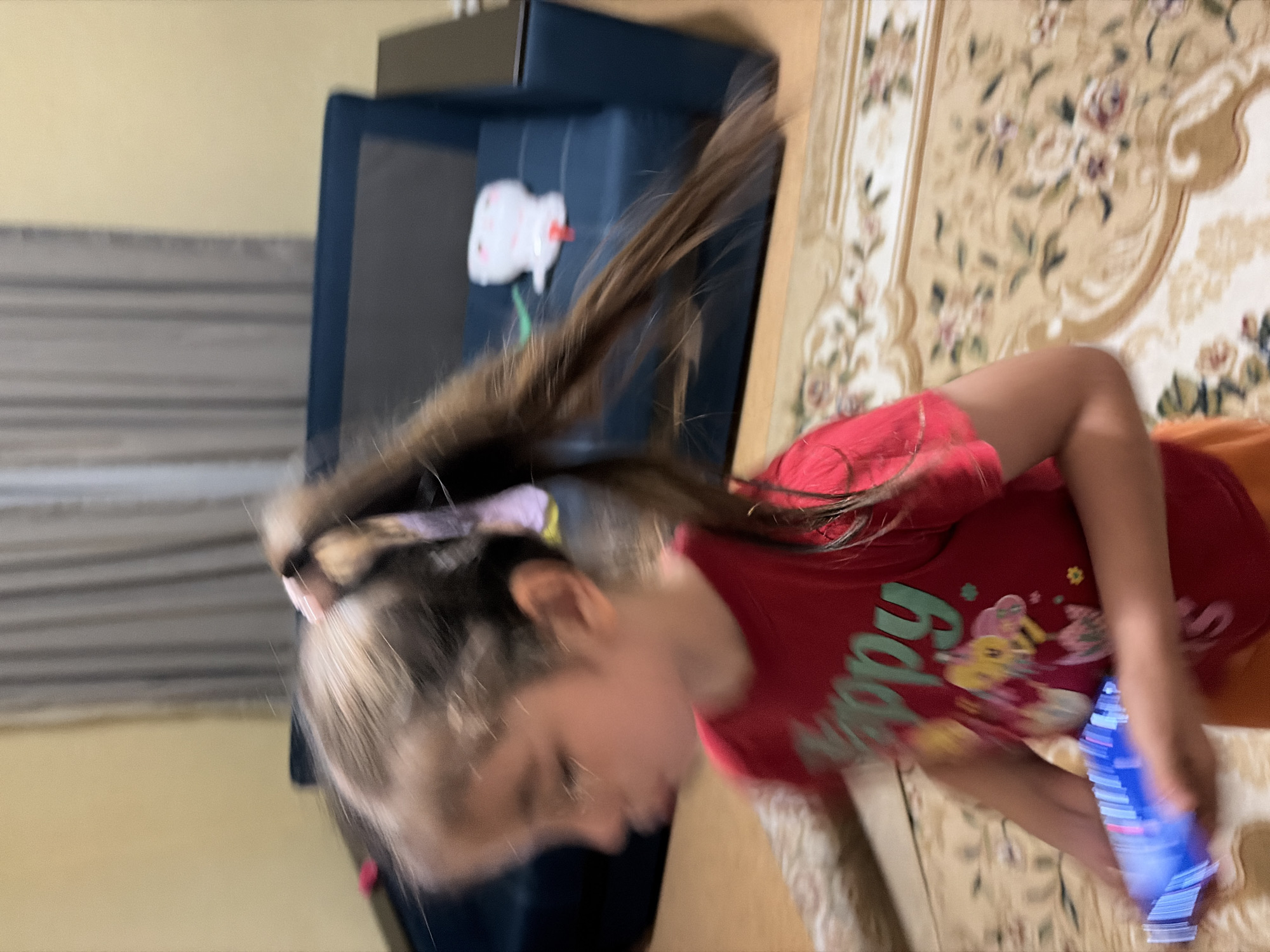 The image shows a young girl with brown hair in a ponytail, wearing a red t-shirt with a graphic design and the word "happy" in green lettering. She is holding a blue object in her hands, which appears to be a small electronic device, possibly a toy or game. The girl is in motion, with the image slightly blurred due to movement.
The background features a dark blue sofa, a light-colored wall, and a patterned carpet on the floor. A white stuffed toy with a red string is visible on the sofa. The overall setting appears to be a room in a residential building.
The image does not contain any specific visual cues to determine its exact location within Tambov, Russia. The context of the image is domestic and ordinary.