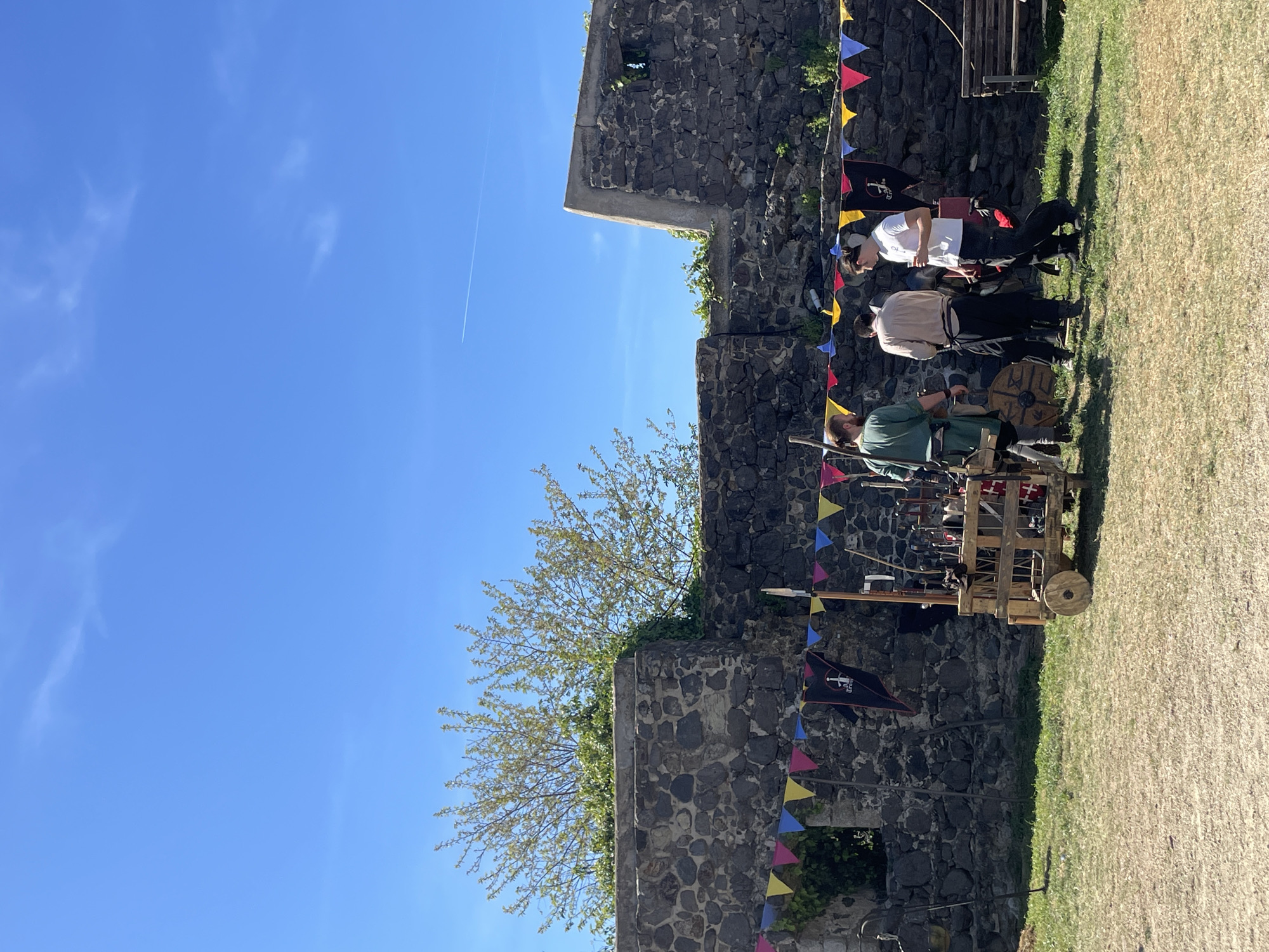 The image shows a section of a stone wall decorated with colorful triangular flags. The wall appears to be part of a larger structure, possibly a castle or fortress. A tree with budding leaves grows near the wall. Three people are positioned near the wall, appearing to be involved in a historical reenactment or display. One person wears a white shirt and dark pants, another wears a light-colored shirt and dark pants, and the third is dressed in green. They are standing near a wooden cart filled with what appears to be historical weapons and equipment. The background includes a clear blue sky with a contrail from an airplane. The scene likely takes place in Hostalric, Spain, based on the presence of a historic fortress or castle and possible reenactment activity.