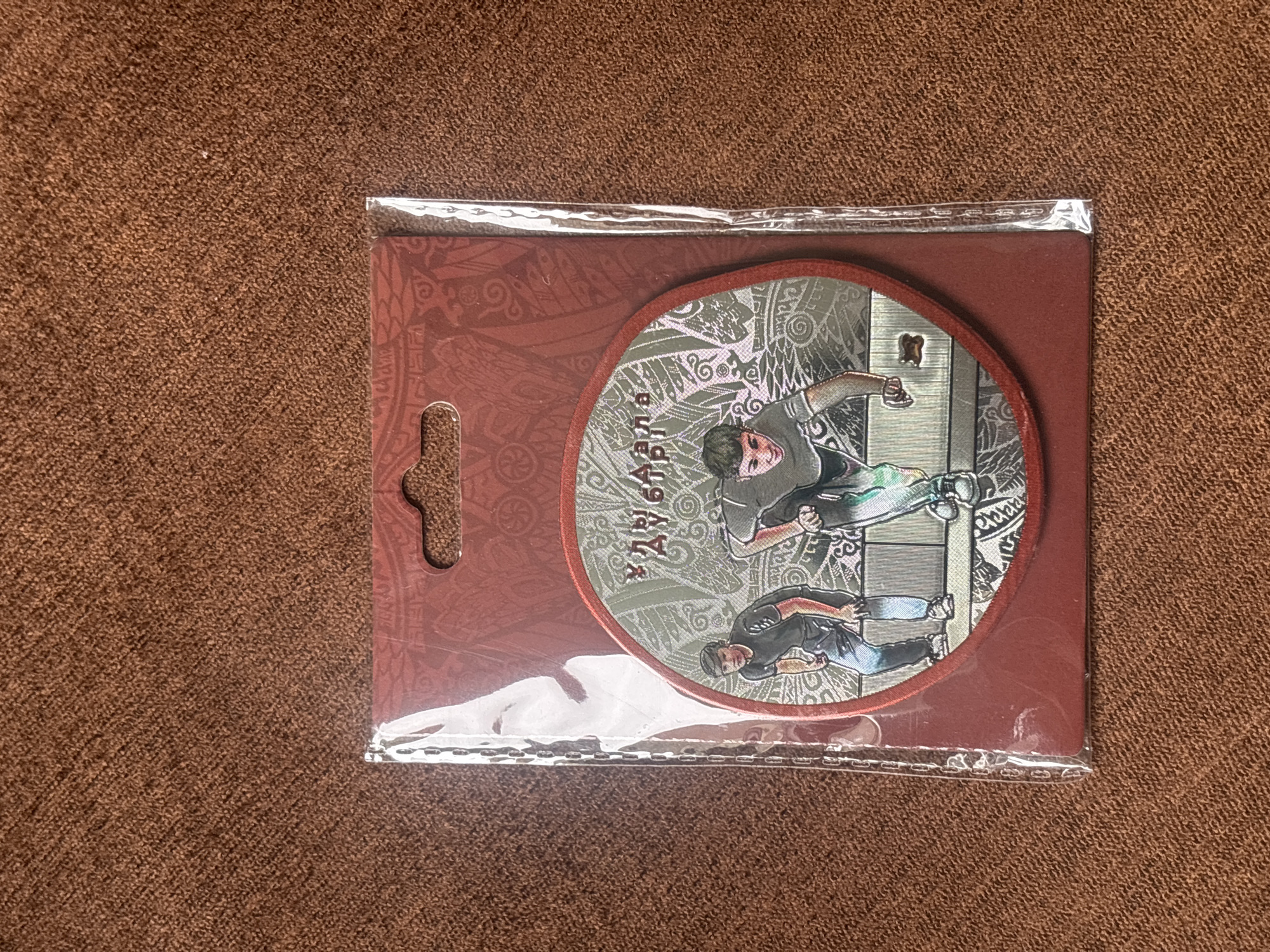 A packaged product is displayed centrally on a textured brown fabric surface. The product consists of a rectangular dark red cardboard backing, featuring subtle patterned ornamentation and a hang tab at the top. This backing encases a circular, multi-layered graphic element, with both components sealed within clear plastic packaging. The circular graphic depicts two young Caucasian males engaged in an urban sport or parkour activity. The male on the left is crouched, wearing a dark t-shirt and light-colored pants, with his hands positioned as if bracing on a low obstacle. The male on the right is in a more active, leaping pose, wearing a dark t-shirt with light accents and dark pants, with his left hand on a grey rectangular barrier. Above the figures, in Cyrillic script, is the text "Улыб ДЛЯ ДУБЛЕРА," which translates to "Smile for the Stuntman." The background of the circular graphic features intricate, light-colored patterns over a grey base, suggestive of urban architectural elements. The scene is static, presented as a product photograph taken in the city of Perm, Russia.