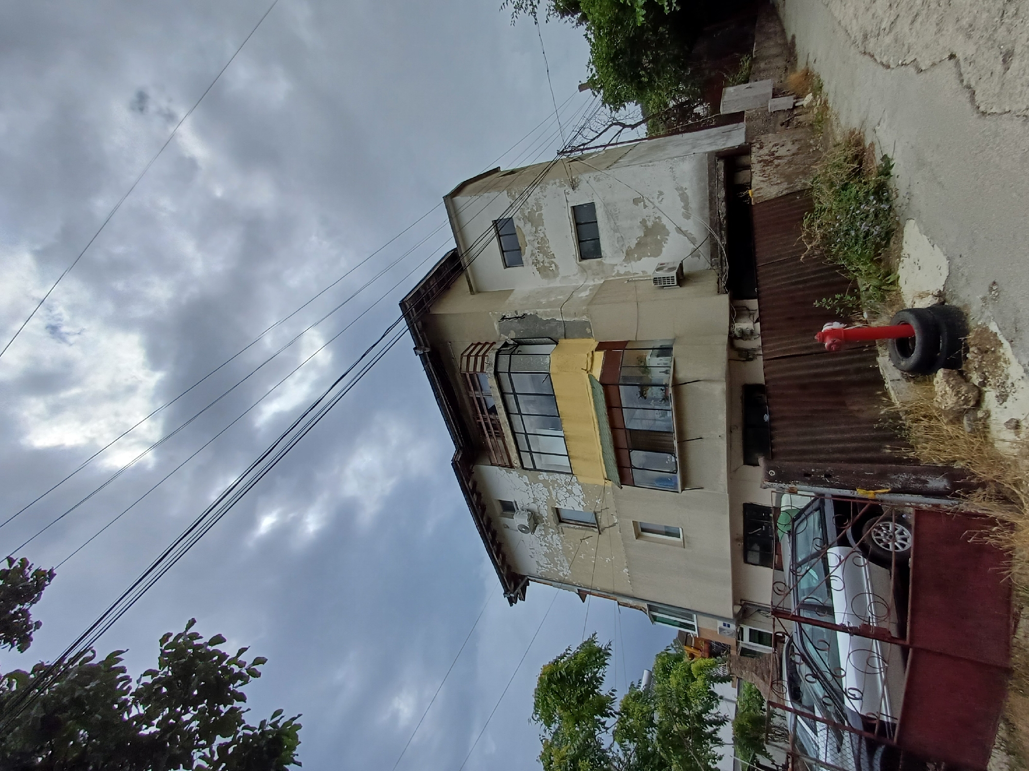 The image captures a side view of a multi-story building in Varna, Bulgaria, under a cloudy sky. The building appears to be weathered, with peeling paint on its facade. Several windows are visible, some featuring balconies with glass enclosures. The ground level is partially obscured by a rusty metal fence and parked cars, including a silver station wagon. Adjacent to the fence, a red fire hydrant stands next to stacked tires. Vegetation, including trees and overgrown bushes, surrounds the building and fence. A paved pathway leads away from the building and the foreground. Power lines are strung across the sky above the building.
