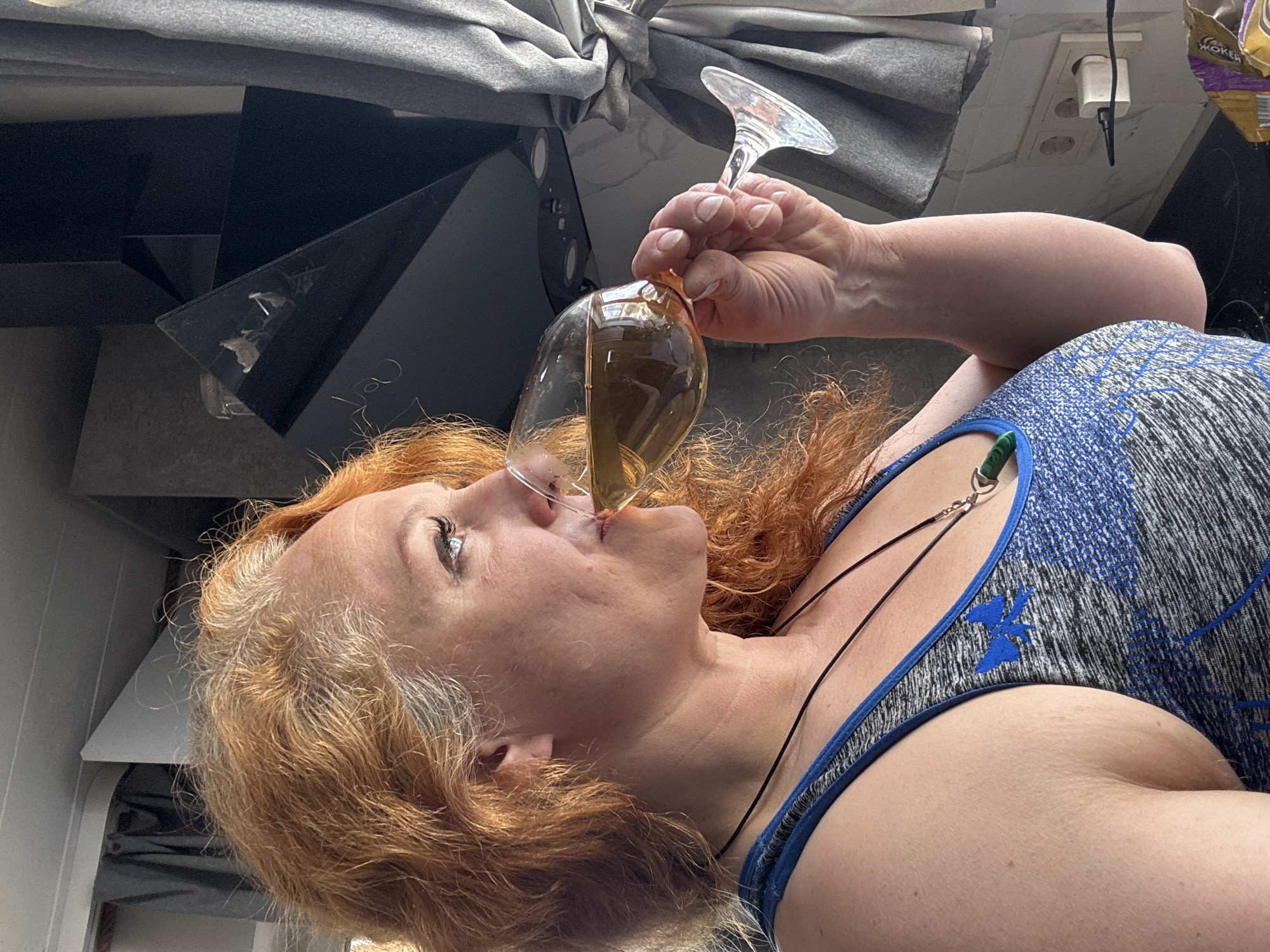 A woman with red hair is drinking from a wine glass. She is wearing a grey and blue tank top. The setting appears to be indoors, potentially in a kitchen, as suggested by the presence of kitchen appliances, a power outlet, and a partially visible bag of cereal. The location is indicated as Chaikovsky, Russia.