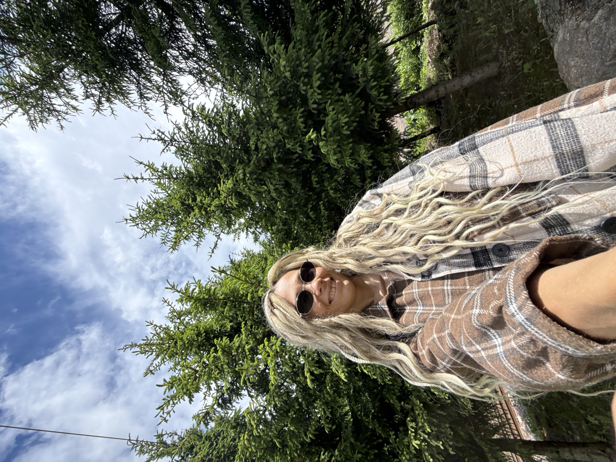 A woman is taking a selfie outdoors, likely in a wooded area. She is wearing sunglasses and a plaid shirt, and has long, curly blonde hair. The background includes evergreen trees and a blue sky with clouds. It's not possible to confirm a location in Rusanovo, Russia from the image alone. There are no indications in the image to prove that this is the location.