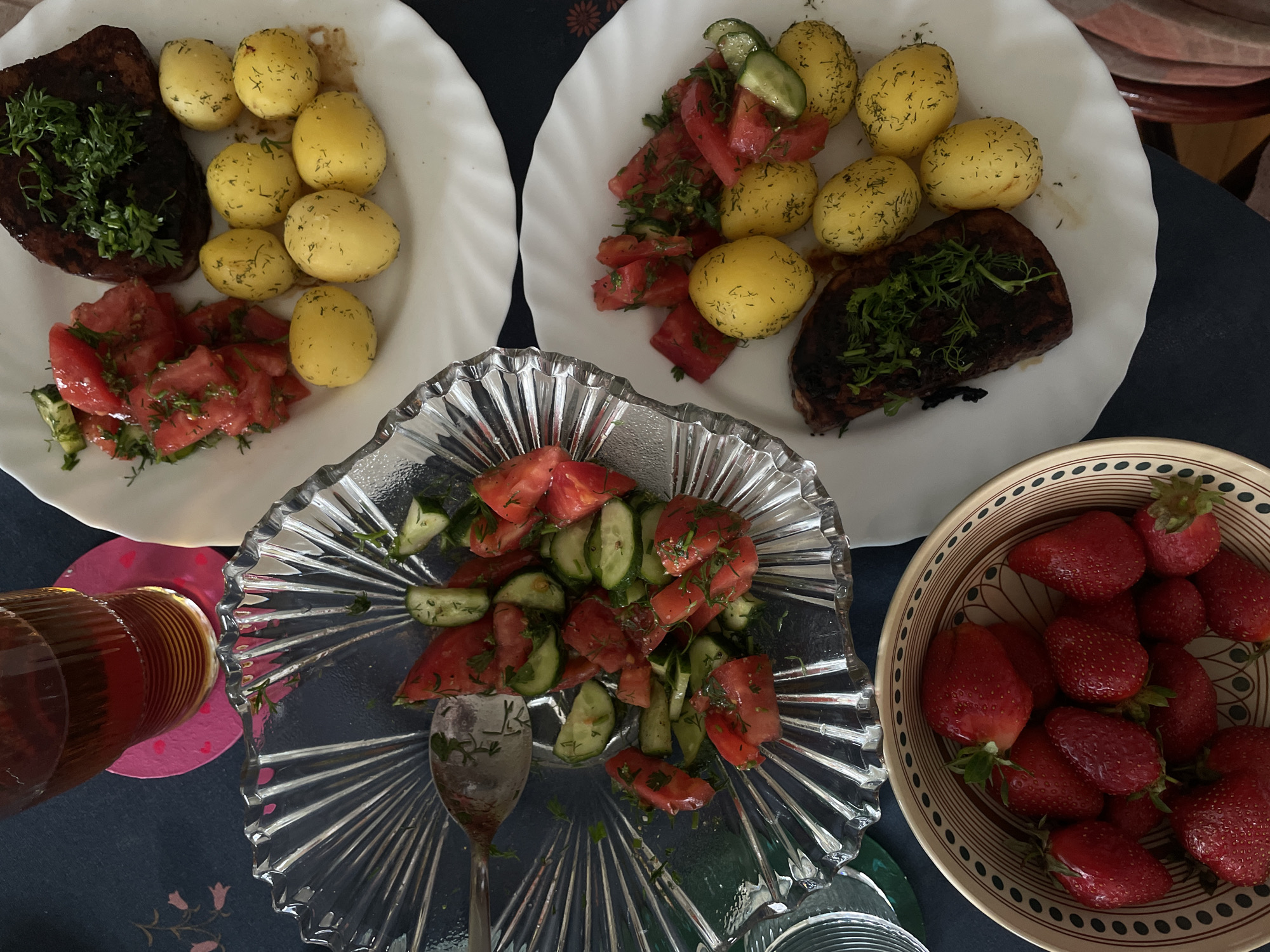 The image depicts a meal consisting of two plates with a serving of meat, potatoes topped with dill, and tomato salad on each. A glass salad bowl containing tomato and cucumber salad stands in the center. A bowl filled with strawberries sits to the right. A glass of a dark beverage is to the left. The setting suggests a casual dining environment. The image does not provide sufficient information to confirm its location in the city of Vracar Urban Municipality, Serbia. No people are visible.