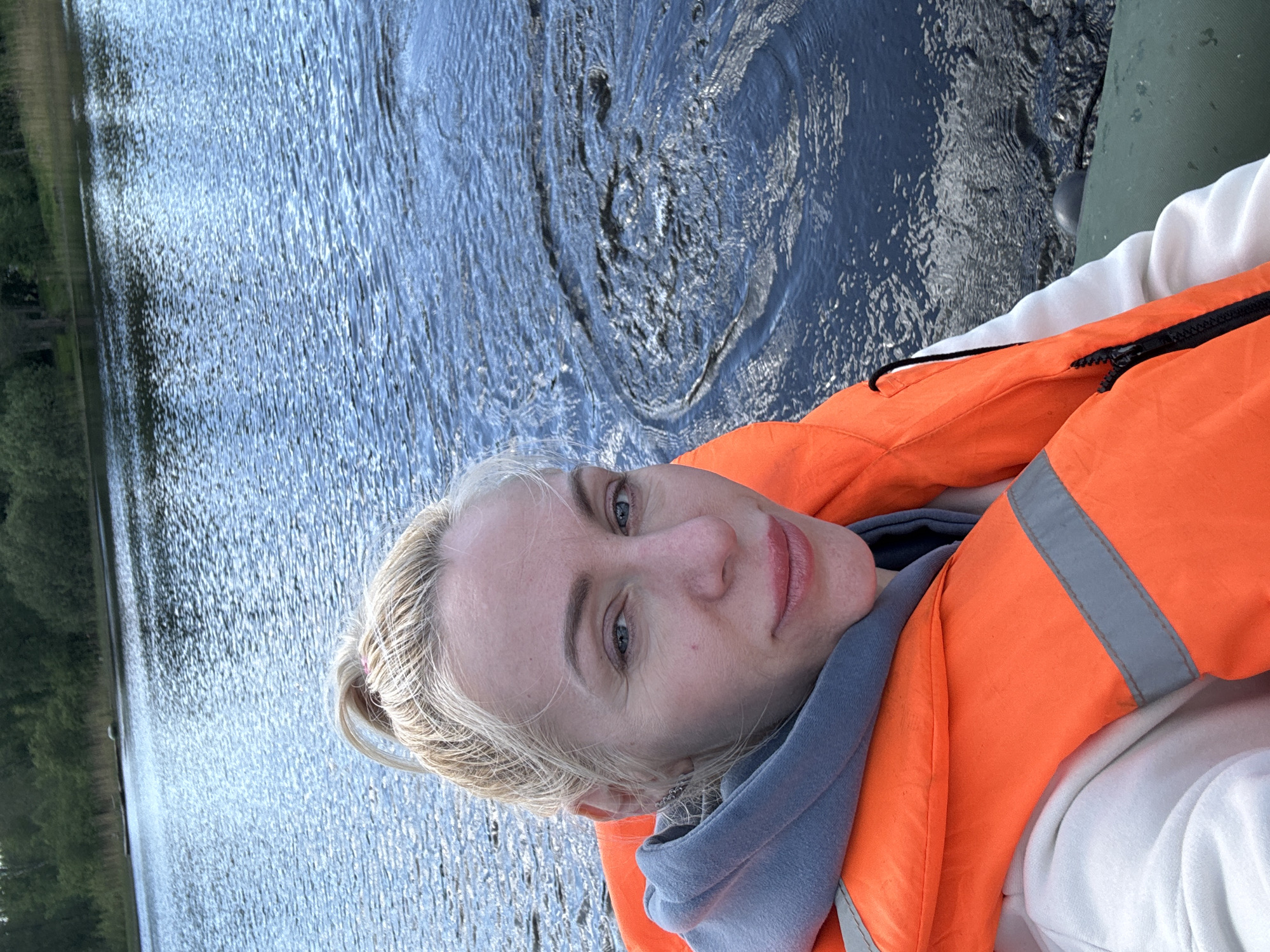 A photograph shows a blonde woman wearing an orange life vest and a grey hoodie over a white shirt. She is on a body of water, possibly a lake or river, with visible water ripples and an adjacent shoreline with trees. The exact location within the city of Unknown, Unknown is impossible to determine from the given image.