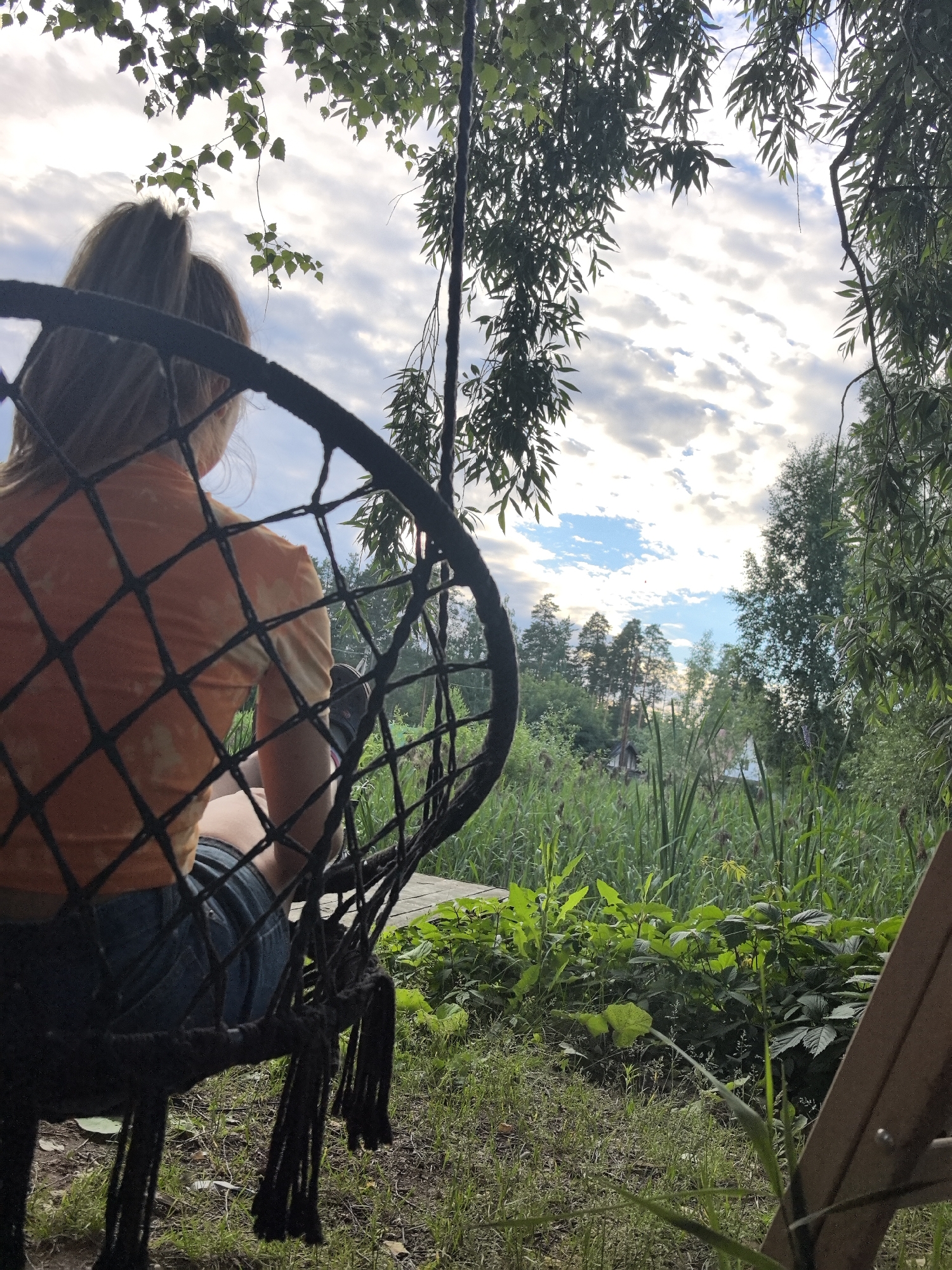 A person sits in a dark, woven hanging chair, viewed from behind. They wear an orange tie-dye t-shirt and denim shorts. The chair is positioned on a wooden platform, with grass and foliage surrounding it. In the background, a lush green landscape unfolds, featuring tall reeds, leafy bushes, and trees. The sky above is partly cloudy, with patches of blue visible through the white clouds. Sunlight filters through the leaves of trees in the foreground, casting dappled shadows. The scene suggests a serene outdoor setting, possibly in a rural or natural area. The overall mood is tranquil and relaxed, with the person appearing contemplative as they gaze out at the scenery. The time of day appears to be late afternoon or early evening, given the soft lighting and the sky conditions. The location context is Noviy, Russia.