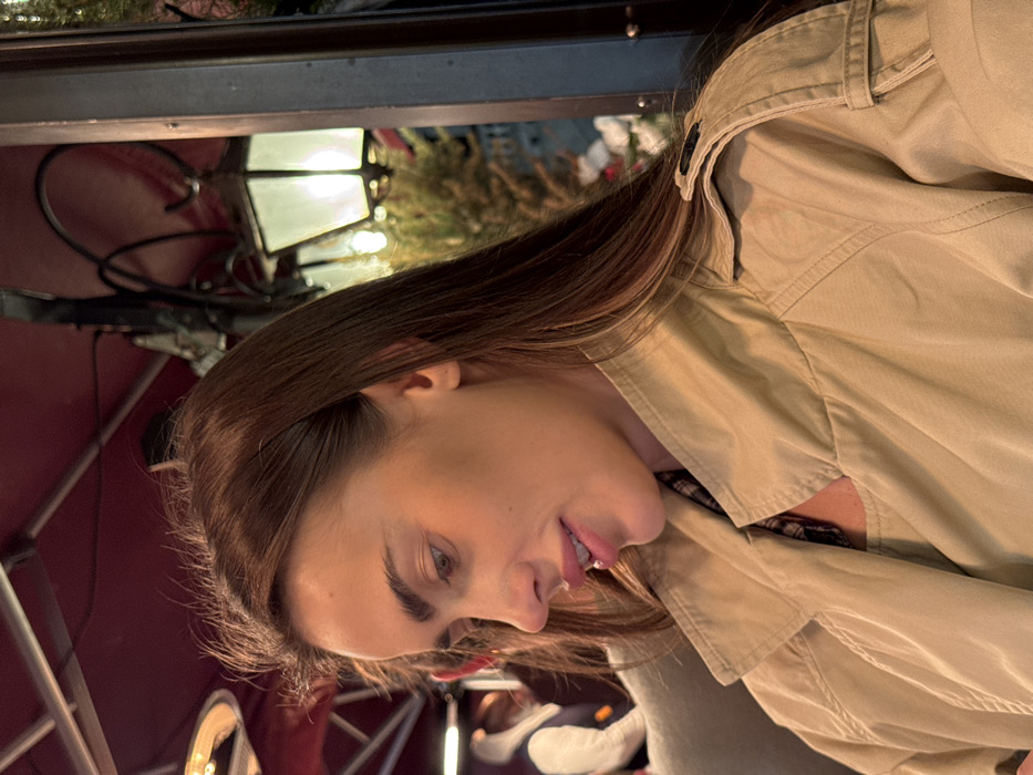 The image features a woman, appearing to be in her late twenties to early thirties, with long, straight, dark brown hair. She is depicted in profile, looking downwards and slightly to her left, exhibiting a subtle, closed-mouth smile. Her facial features include light brown eyes, prominent eyebrows, and full lips. She is wearing a light tan or beige trench coat, with a glimpse of a dark, patterned garment visible beneath the collar. The scene is set in an outdoor or semi-outdoor seating area in Venice, Italy, characterized by a red overhead awning and a ceiling-mounted, circular radiant heater in the upper left. In the background, a decorative black metal street lamp with a glowing white light source is present on the right, alongside blurred green foliage. A partially visible figure in a light-colored hooded jacket is discernible in the far left background. The ambient lighting suggests an evening or nighttime environment.